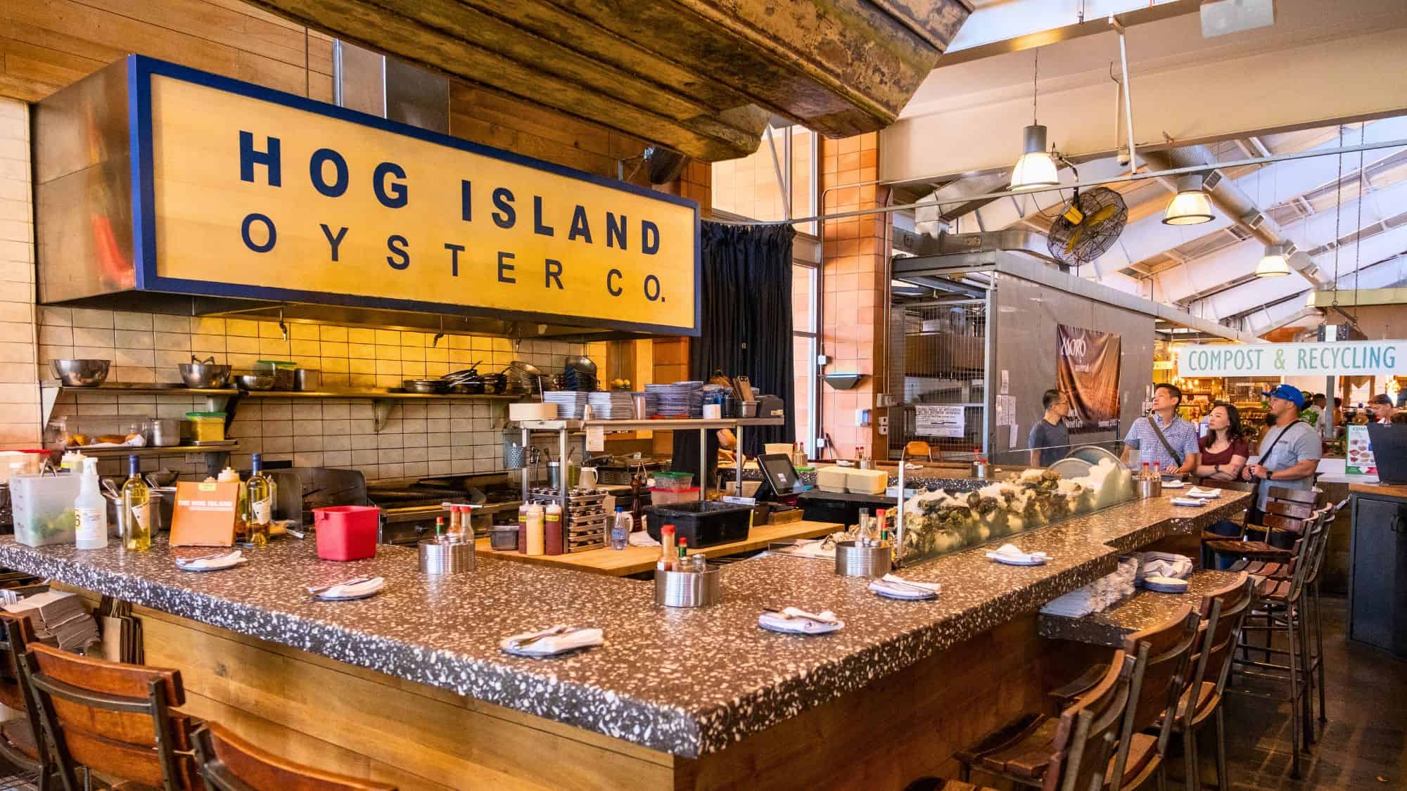 The interior of Hog Island Oyster Co., an oyster bar with a rustic, seafood-focused atmosphere. The long counter features a large yellow and blue sign, fresh oysters on ice, and condiments for customers to enjoy, with a lively open kitchen in the background.