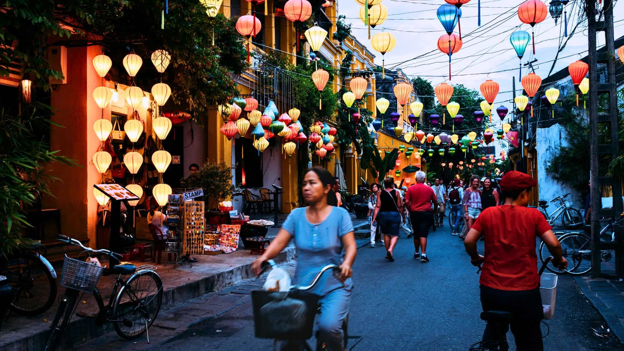 A vibrant night market scene in Hoi An, with colorful lanterns hanging overhead, illuminating the bustling street. Locals and tourists walk or ride bicycles past market stalls and cafes, soaking in the warm glow of this enchanting ancient town.