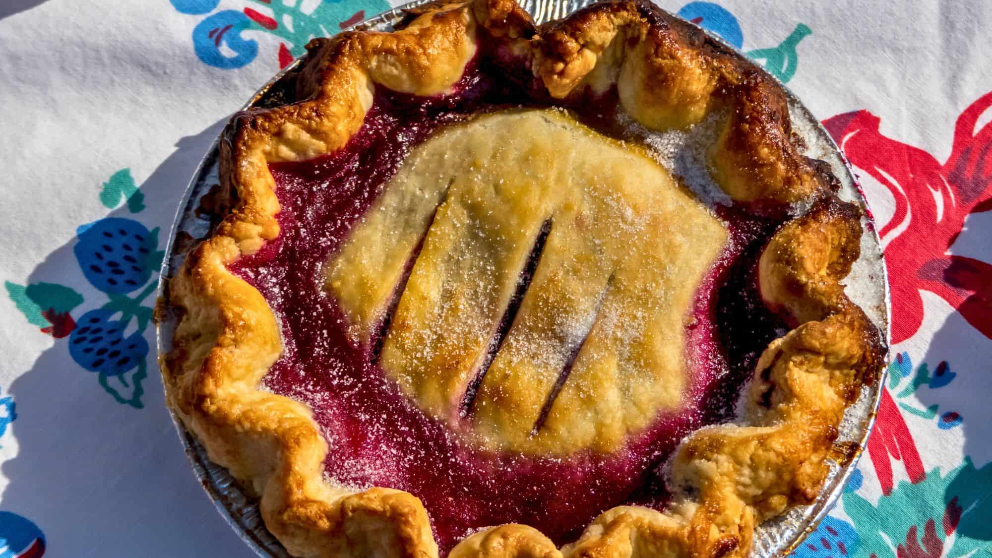 A homemade huckleberry pie with a golden, flaky crust and a deep purple berry filling peeking through slits in the top. The pie sits on a colorful tablecloth with a berry pattern, dusted with sugar for a finishing touch.