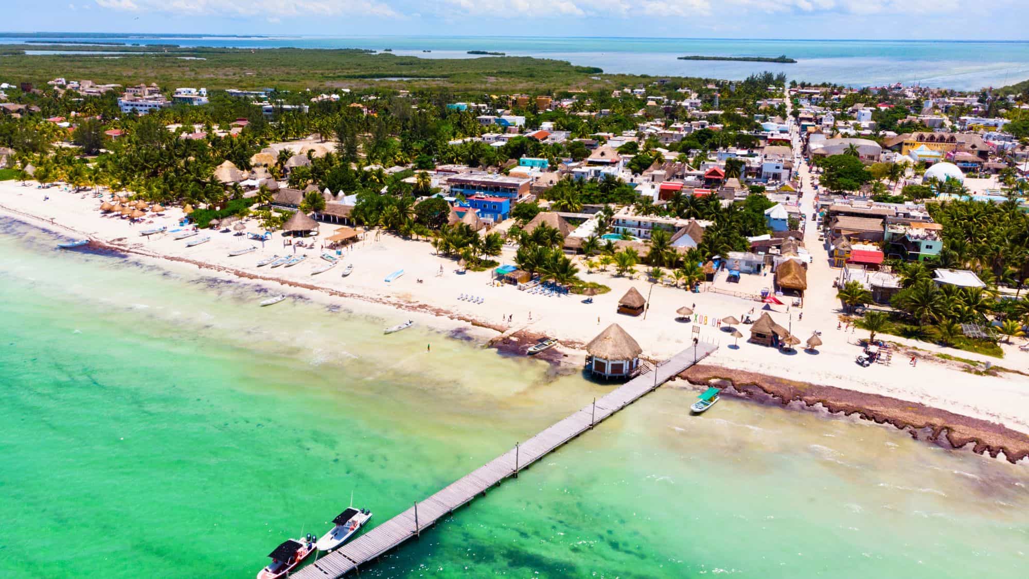 Aerial view of a tropical beach town with palm trees, colorful buildings, and white sandy shores. A long wooden pier extends into the shallow, turquoise waters, with boats anchored nearby.