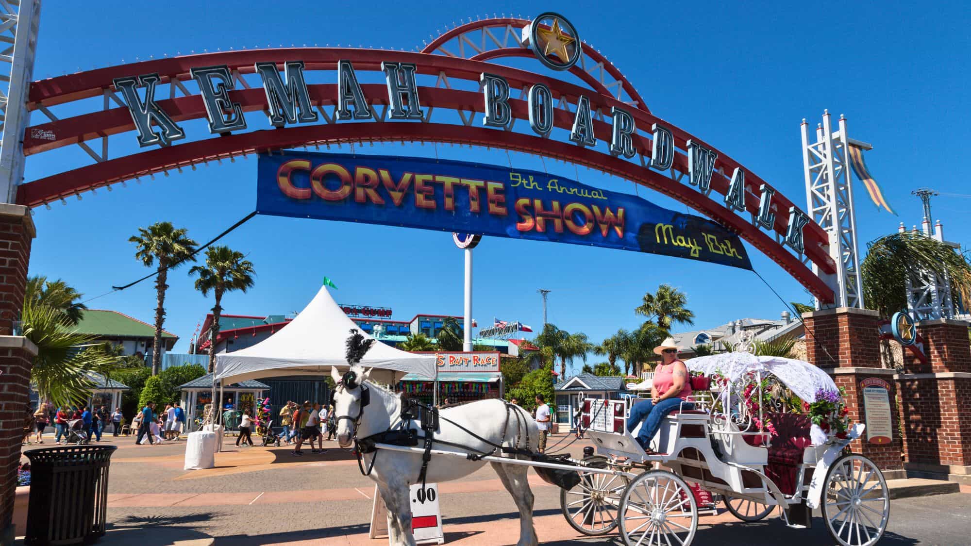 A lively scene at the entrance of Kemah Boardwalk, Texas, featuring a red and white archway with "Kemah Boardwalk" written on it. A horse-drawn carriage with white decor and a female driver adds a nostalgic charm to the bustling amusement area.