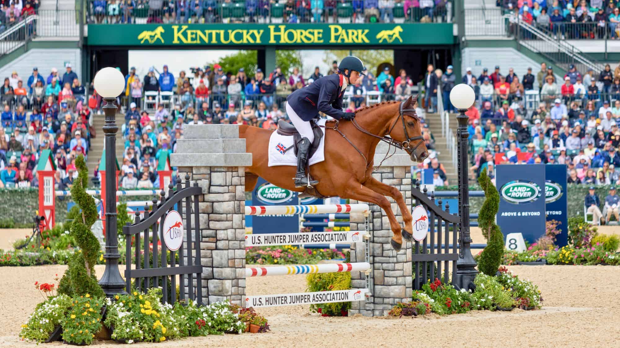 A professional equestrian in a navy jacket and helmet rides a brown horse mid-jump over a U.S. Hunter Jumper Association obstacle at Kentucky Horse Park. A crowd watches in the background during a prestigious horse-jumping event.