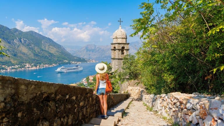A woman in a white tank top and denim shorts walks down a historic stone pathway in Kotor, Montenegro, with a scenic view of the bay, cruise ships, and an old church tower.