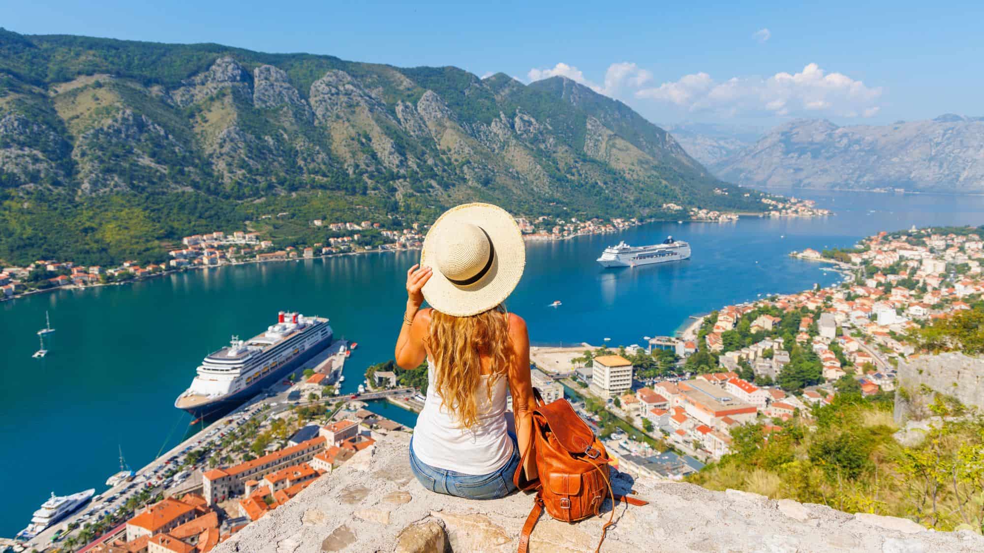 A woman in a white tank top and straw hat sits on a rocky ledge overlooking the stunning Bay of Kotor, with cruise ships docked at the waterfront and lush mountains in the background.