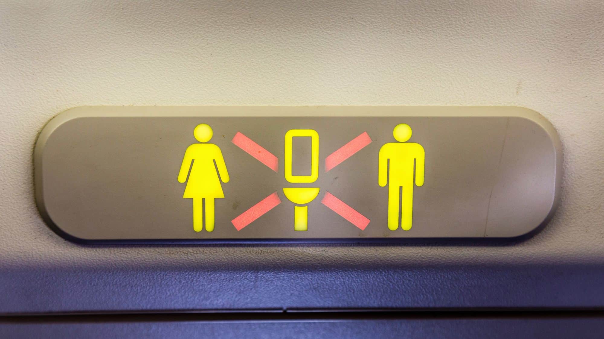 An airplane lavatory sign shows a red "X" over a toilet symbol, flanked by illuminated icons of a man and woman, indicating the restroom is occupied.