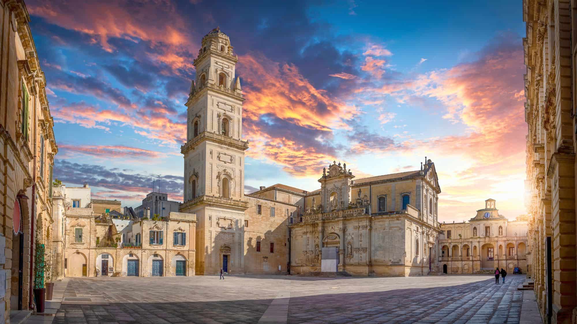 The historic baroque architecture of Lecce, Italy, is bathed in the warm hues of a sunset, highlighting the intricate details of the cathedral and the surrounding square.