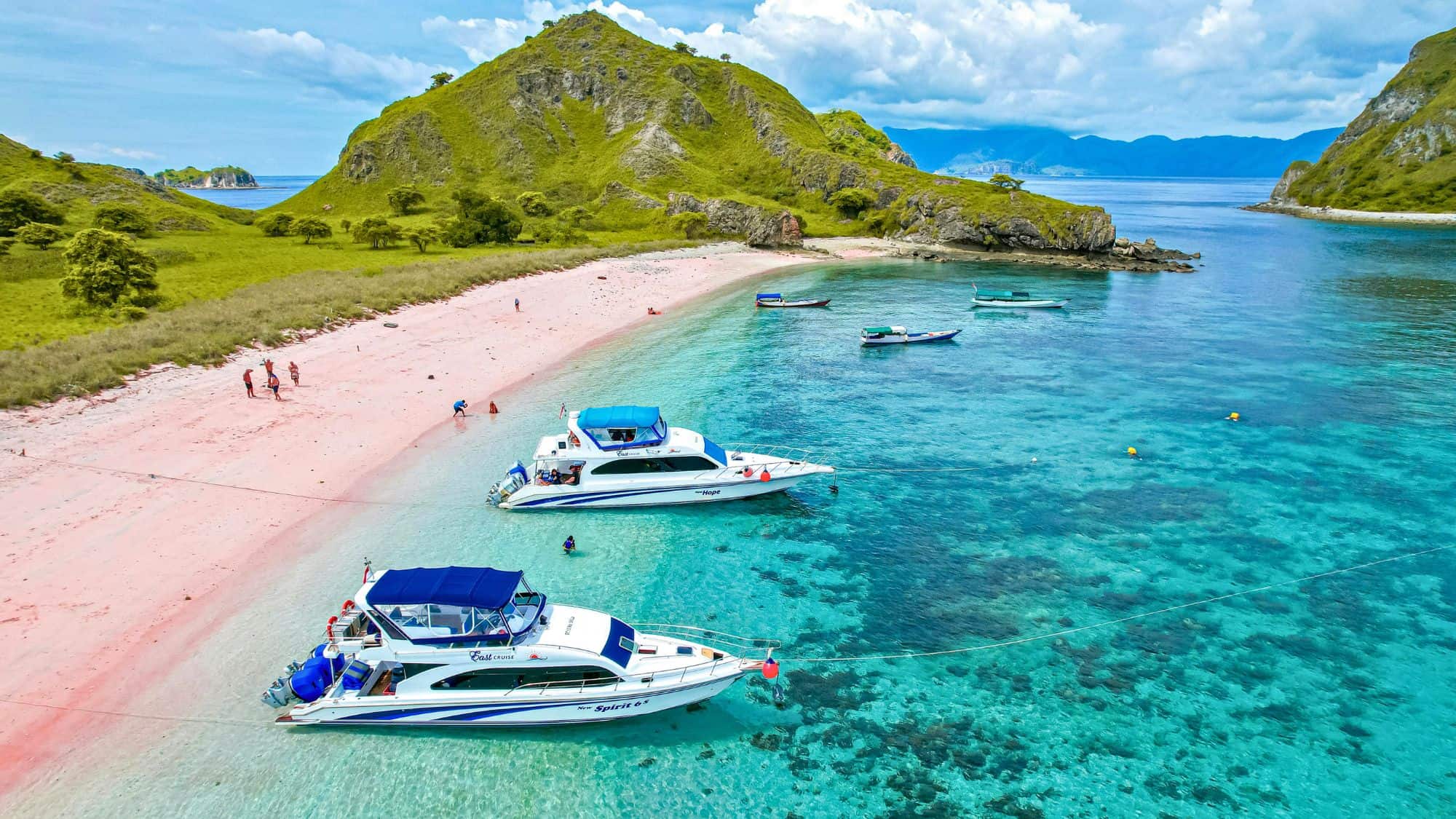 Crystal-clear turquoise waters lap against a pink sand beach in Lombok, Indonesia, where speedboats are docked near green, rolling hills.