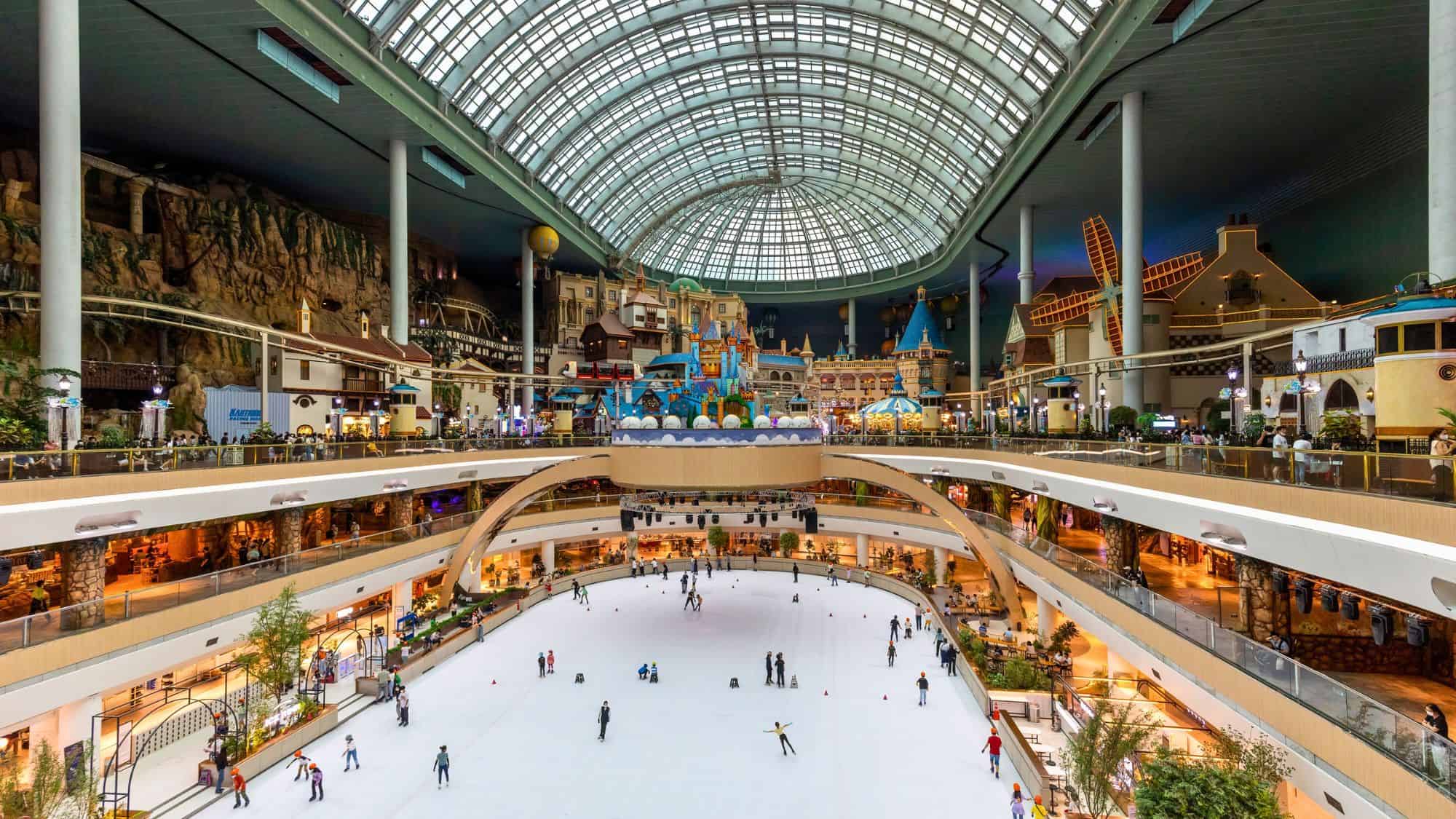 A massive indoor ice-skating rink at Lotte World in South Korea, surrounded by multiple levels of attractions, European-style facades, and a glass-domed ceiling.