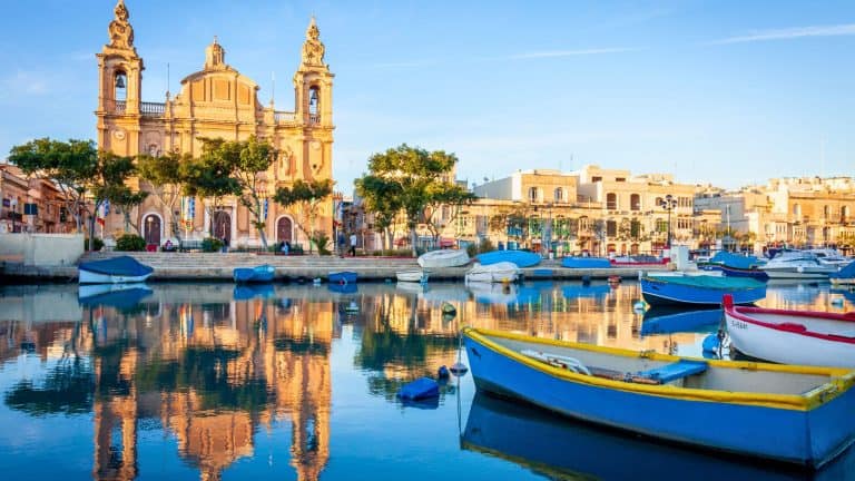 Charming harbor scene with colorful wooden fishing boats bobbing in calm blue water, set against a backdrop of historic buildings and a stately church in a Maltese village.