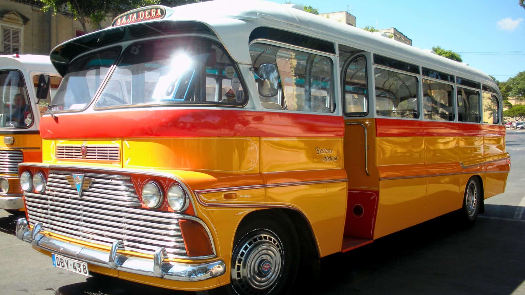 Brightly colored vintage Maltese bus with bold yellow, red, and orange paint parked in a sunny town square, exuding retro charm and local character.