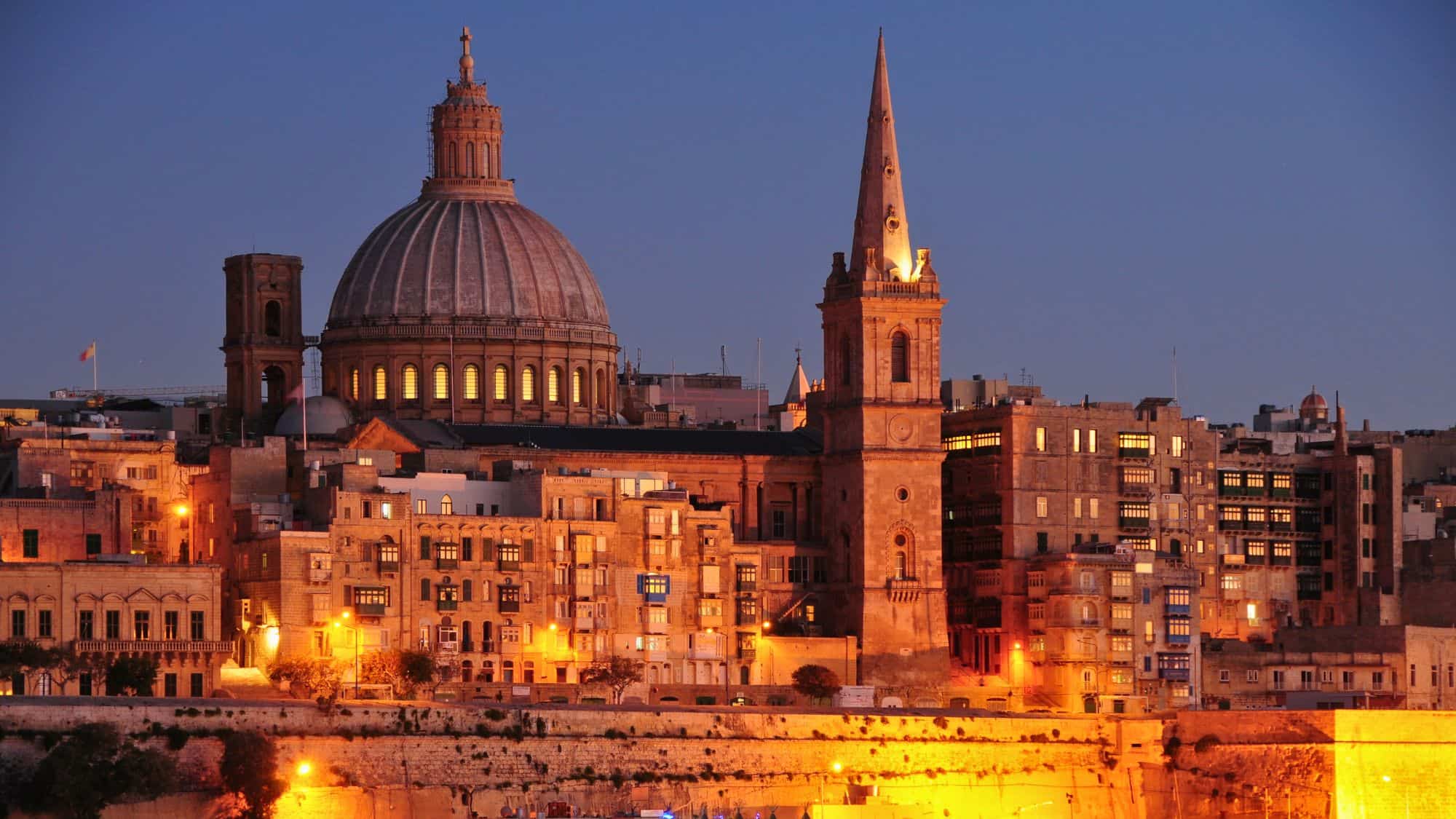 The lit-up dome and spire of a grand Baroque church in Valletta, Malta, glow warmly against the deep blue twilight sky, surrounded by historic limestone buildings.