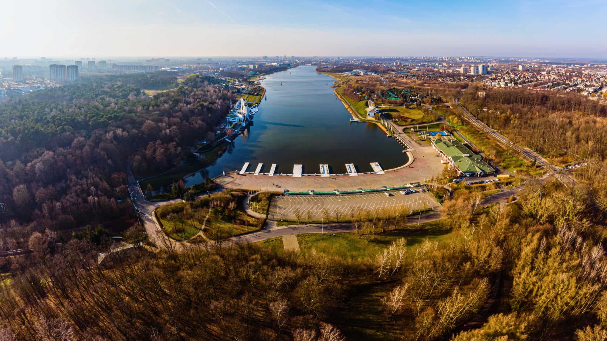Expansive aerial view of a rowing and recreation center on a long man-made lake, with docks, boathouses, and a tree-lined park set against a city skyline in the distance.