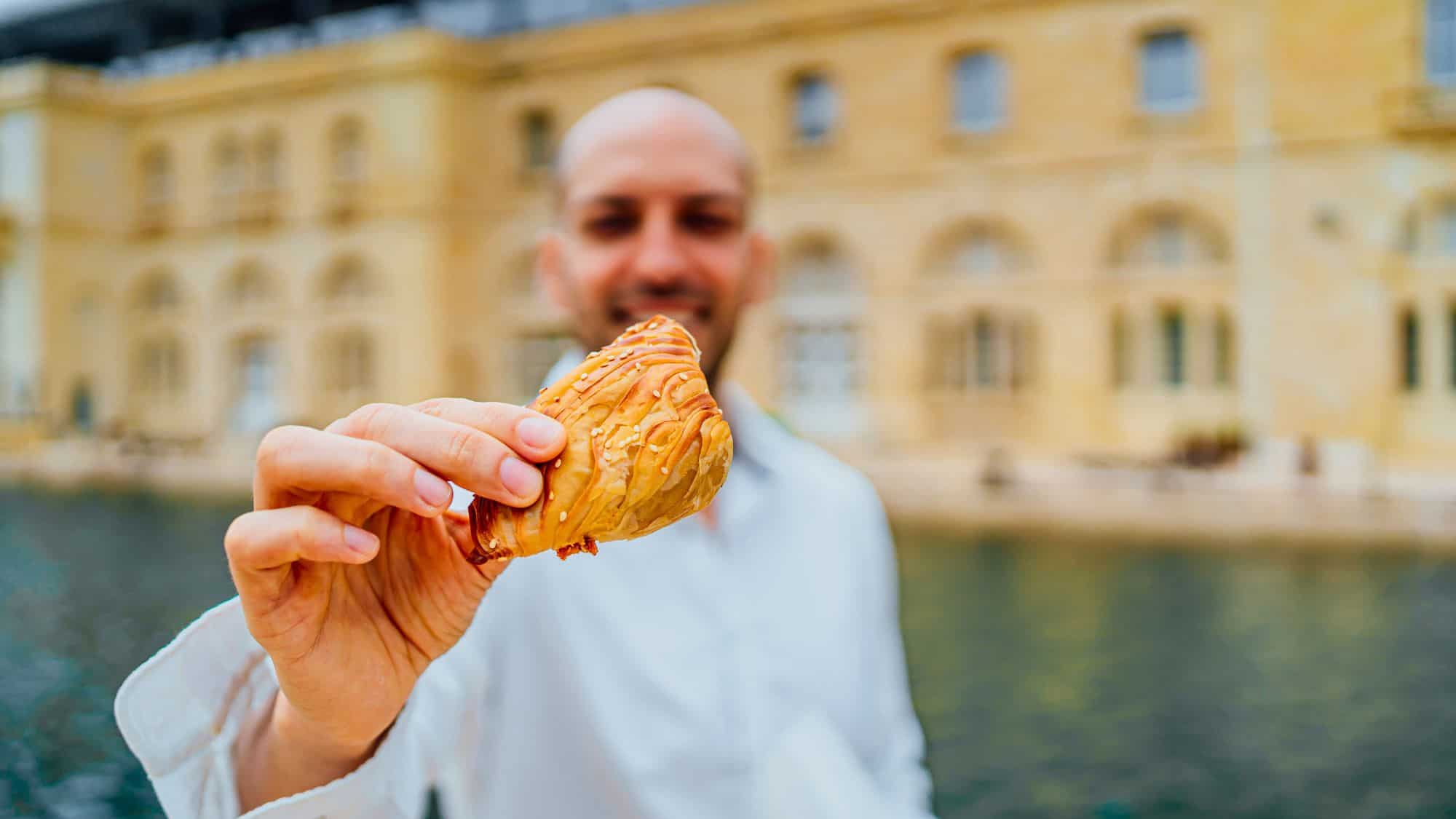 Close-up of a person holding a traditional flaky Maltese pastry, with sesame seeds on top, in focus while the background shows a limestone building by the water.