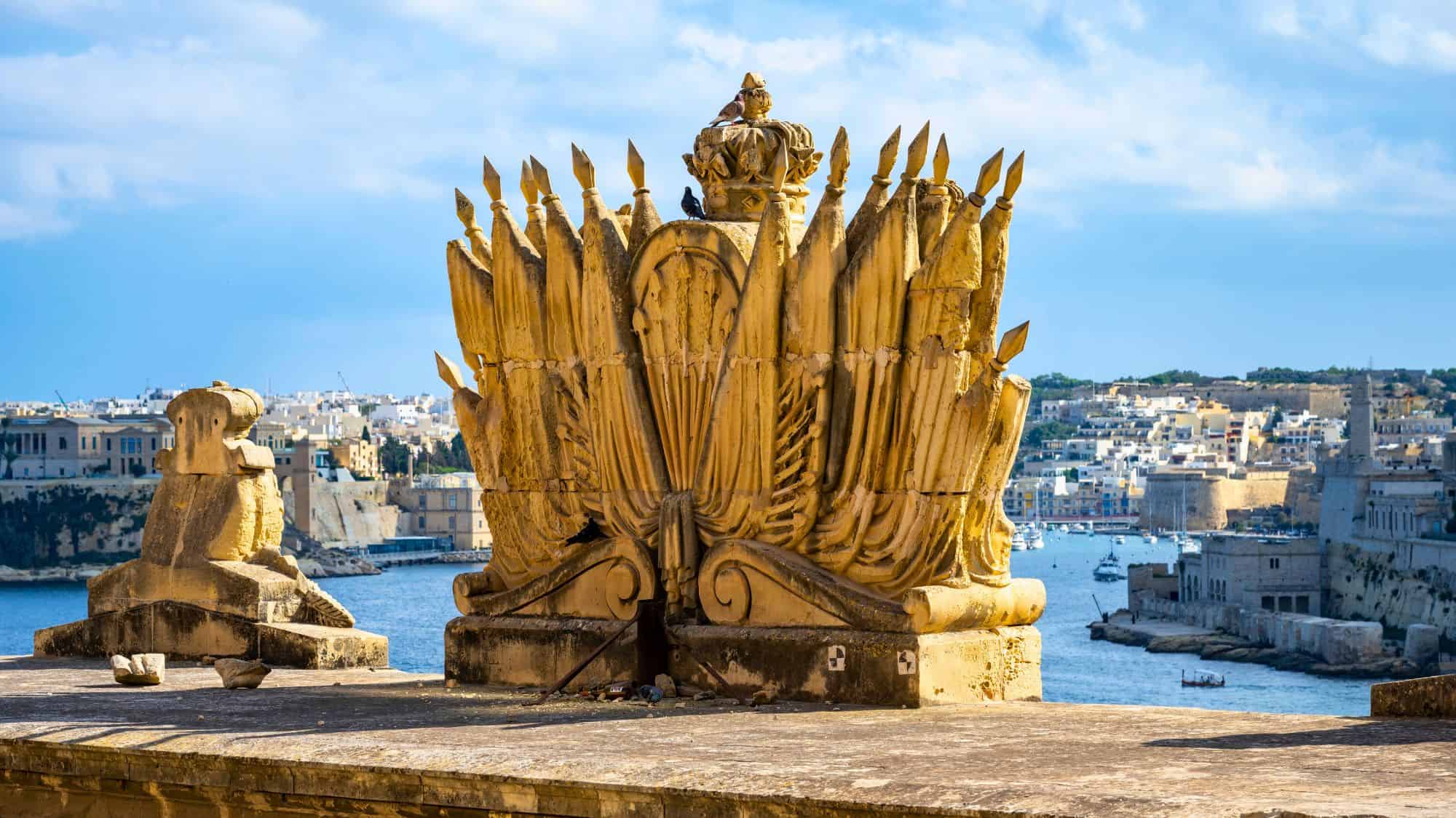 Ornate stone sculpture of a Maltese noble crest, adorned with spears and feathers, on a rooftop overlooking a historic harbor and stone city across the water.