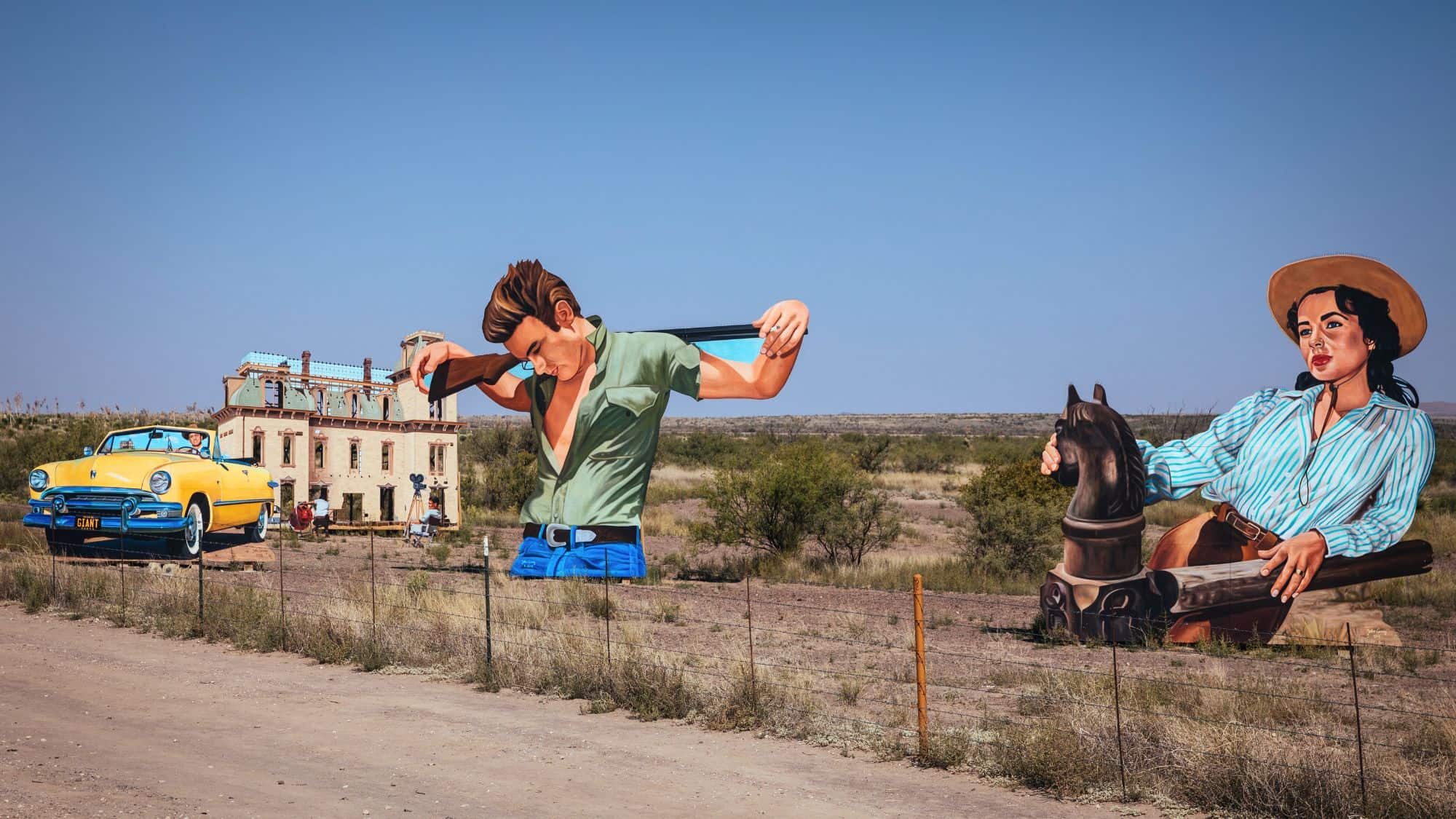 An art installation in the desert near Marfa, Texas features oversized cutouts of 1950s movie characters, a yellow convertible, and a film set house, set against a stark blue sky.