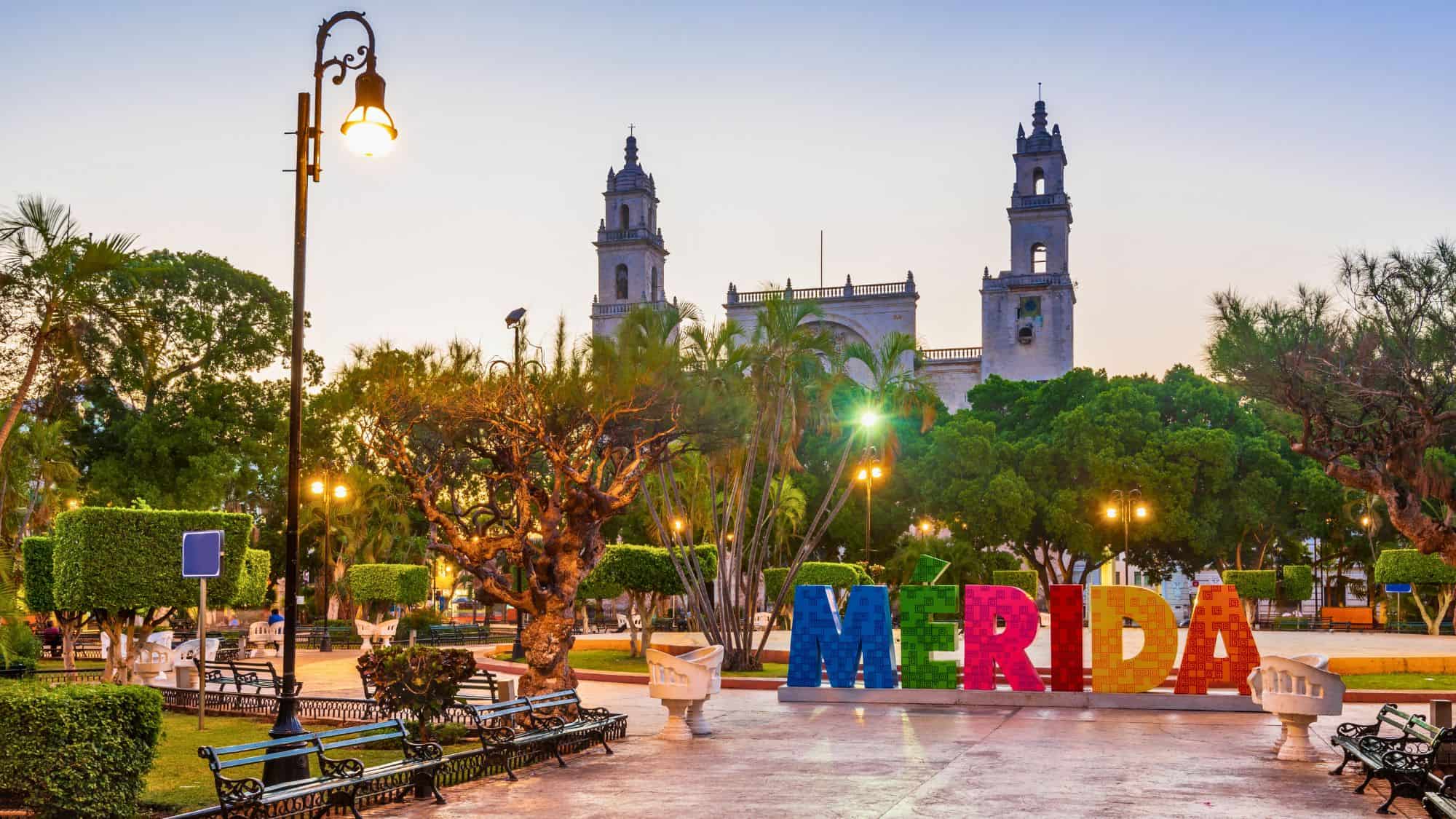 A charming park in Merida, Mexico, is illuminated at dusk, featuring the grand Cathedral of Mérida in the background and the vibrant "Mérida" sign in the foreground.