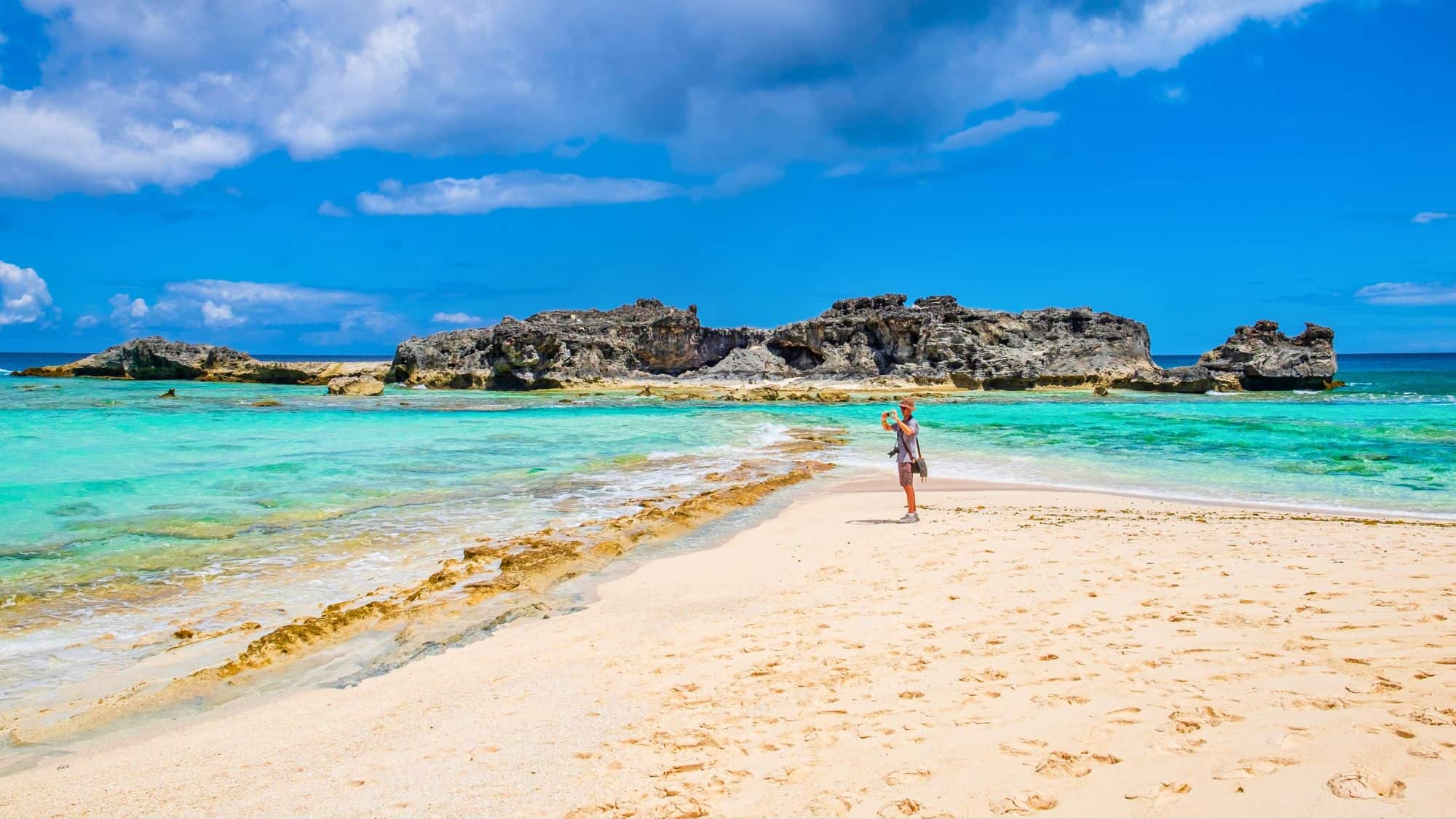 A stunning beach with clear, shallow waters and rugged rock formations in the distance. A person stands on the soft, golden sand, taking in the breathtaking ocean view.
