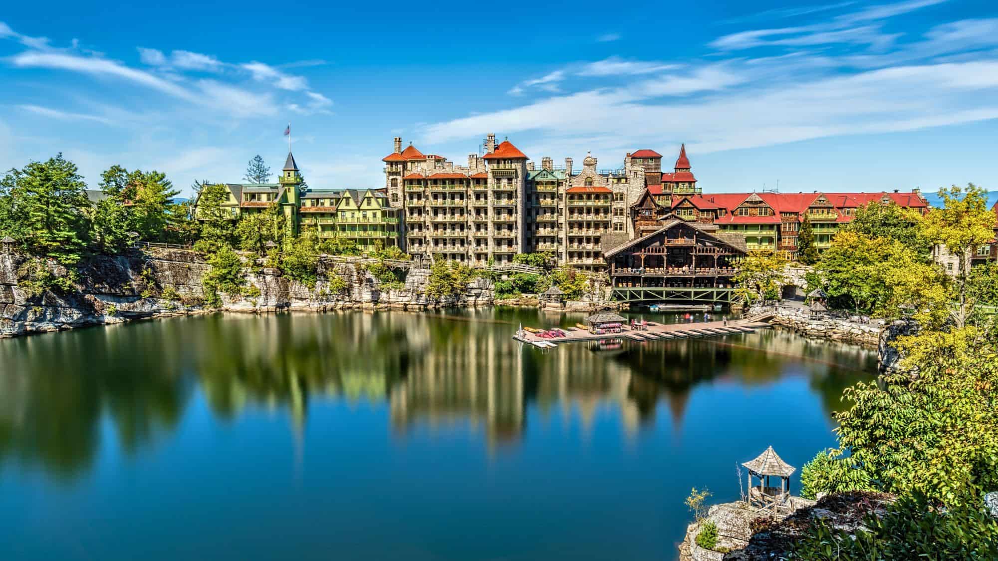 A grand historic resort with red and green roofs sitting beside a reflective lake, surrounded by cliffs and lush trees. The calm water mirrors the building, creating a stunning, tranquil view.