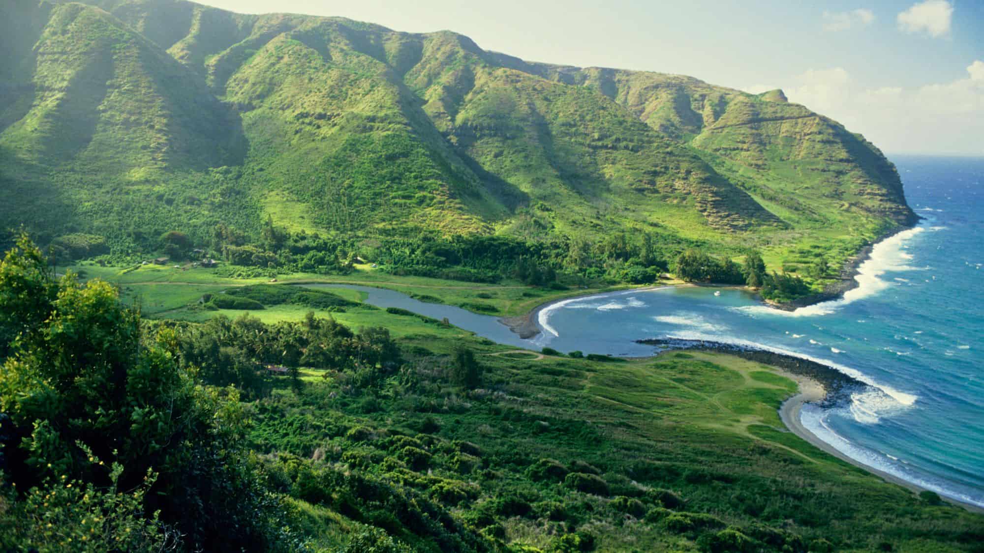 A remote green valley on Molokai, Hawaii leads to a crescent-shaped beach with turquoise waters and dramatic ridgelines, exuding untouched tropical beauty.