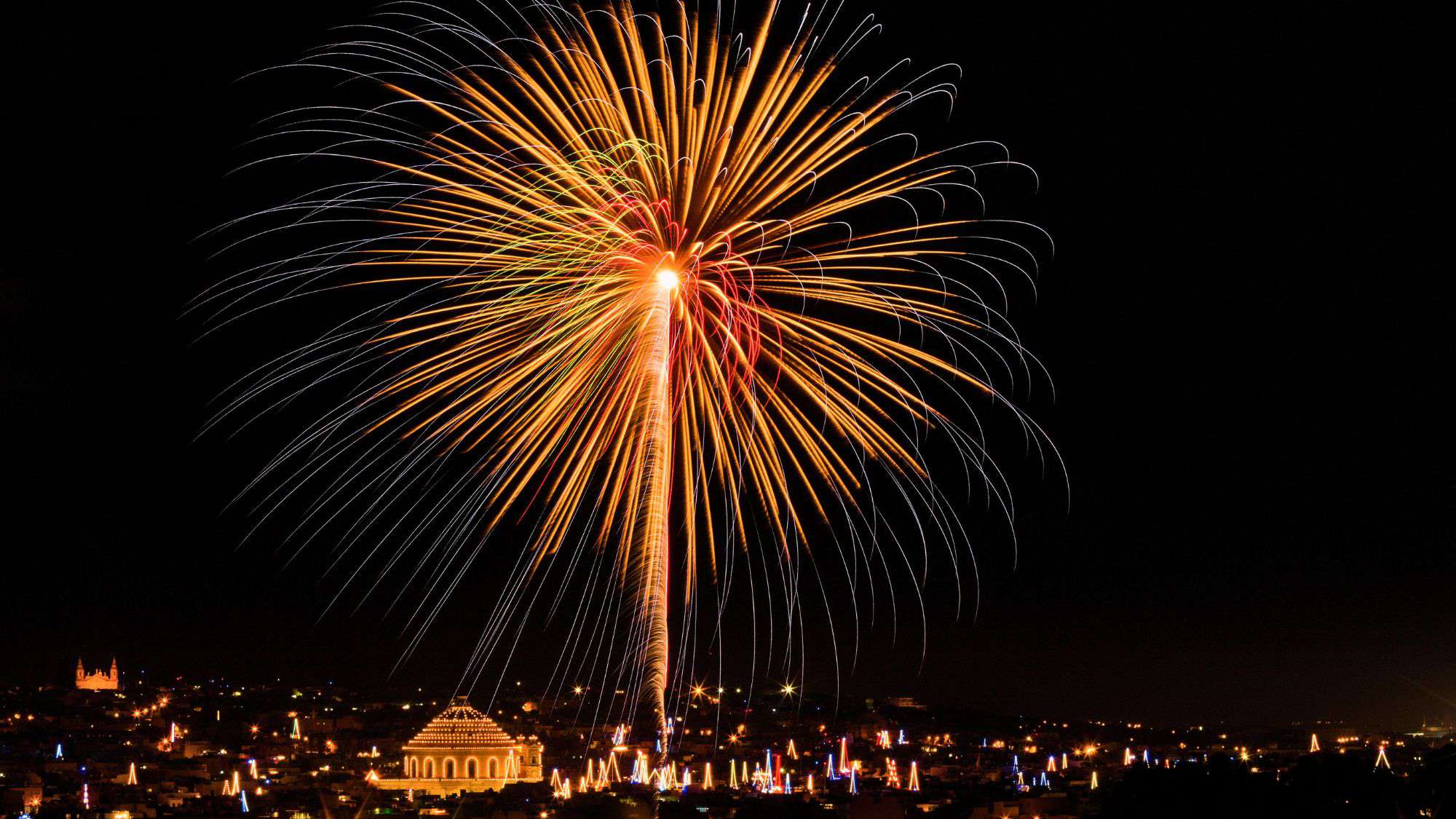Spectacular burst of golden and red fireworks lighting up the night sky over a Maltese town, with the iconic dome of Mosta’s church illuminated below.