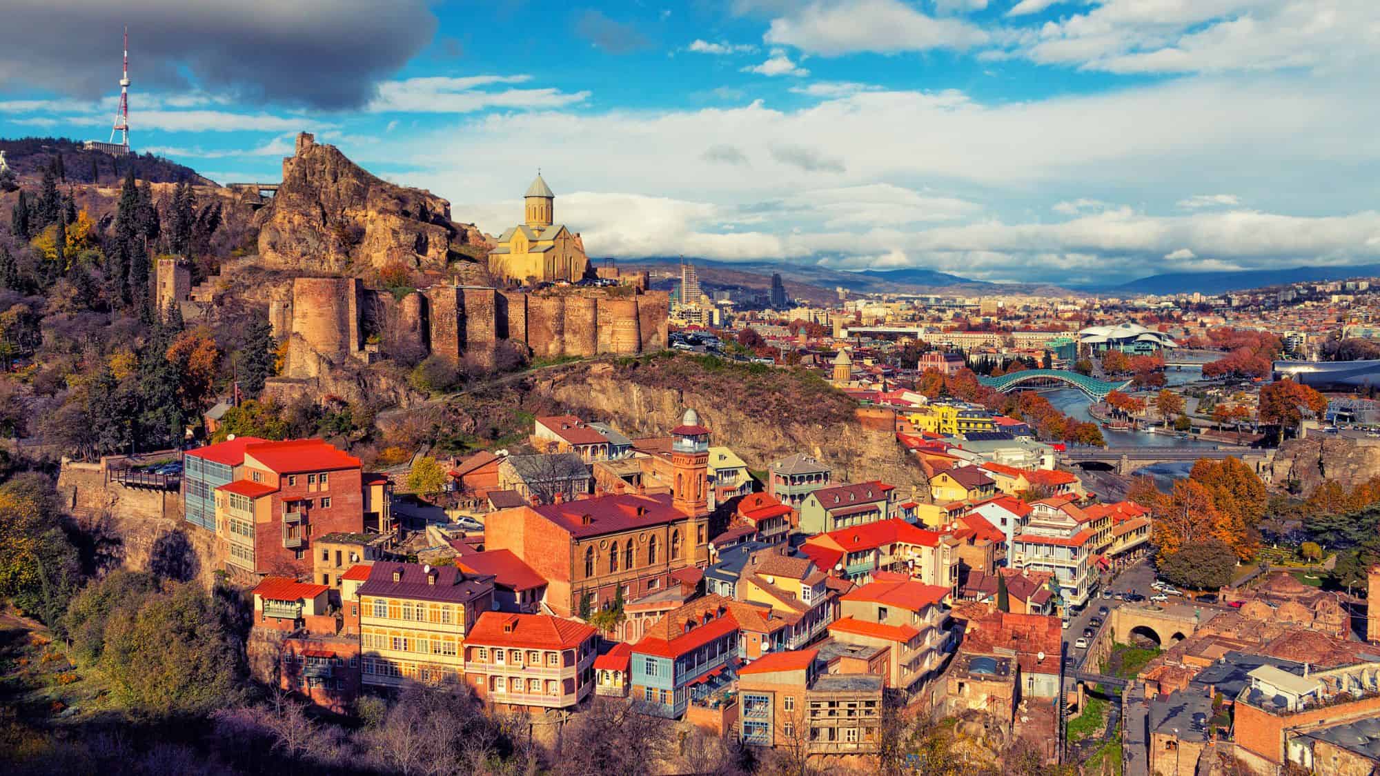 A panoramic view of Tbilisi, Georgia, featuring the ancient Narikala Fortress atop a rocky hill, surrounded by colorful houses and the modern cityscape.