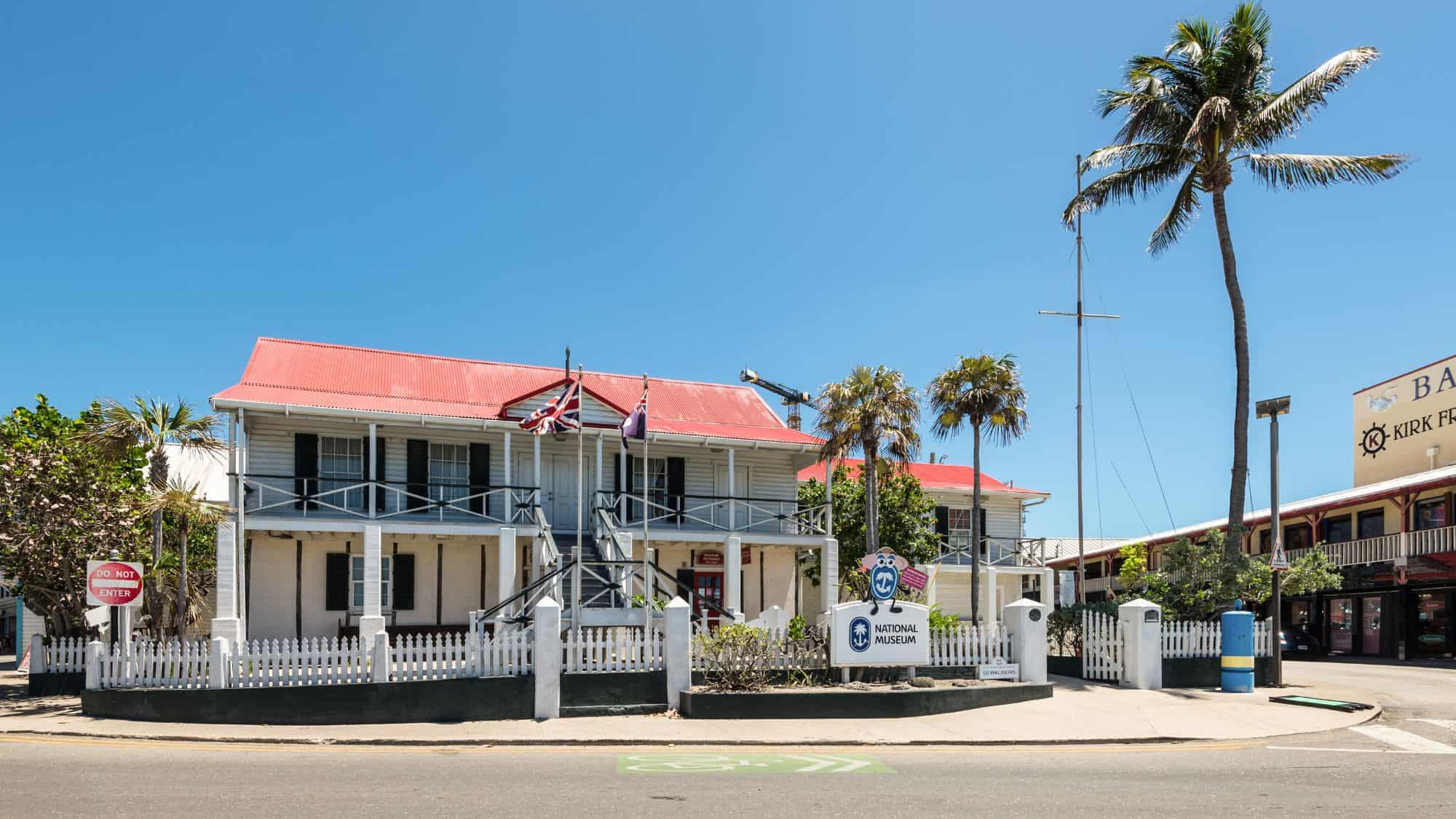 A historic colonial-style building with a red roof and white picket fence, housing the National Gallery. The Union Jack and Cayman Islands flags wave in front, marking it as a cultural landmark.