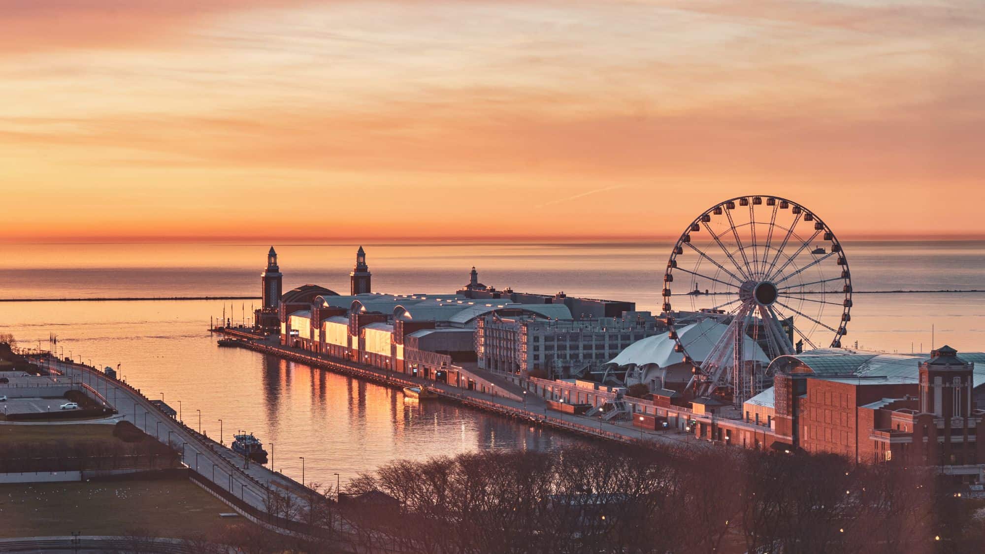 A scenic aerial view of Navy Pier in Chicago during sunset, with the iconic Centennial Wheel standing tall against the golden sky. The pier’s buildings and Lake Michigan’s calm waters reflect the warm hues of the setting sun.