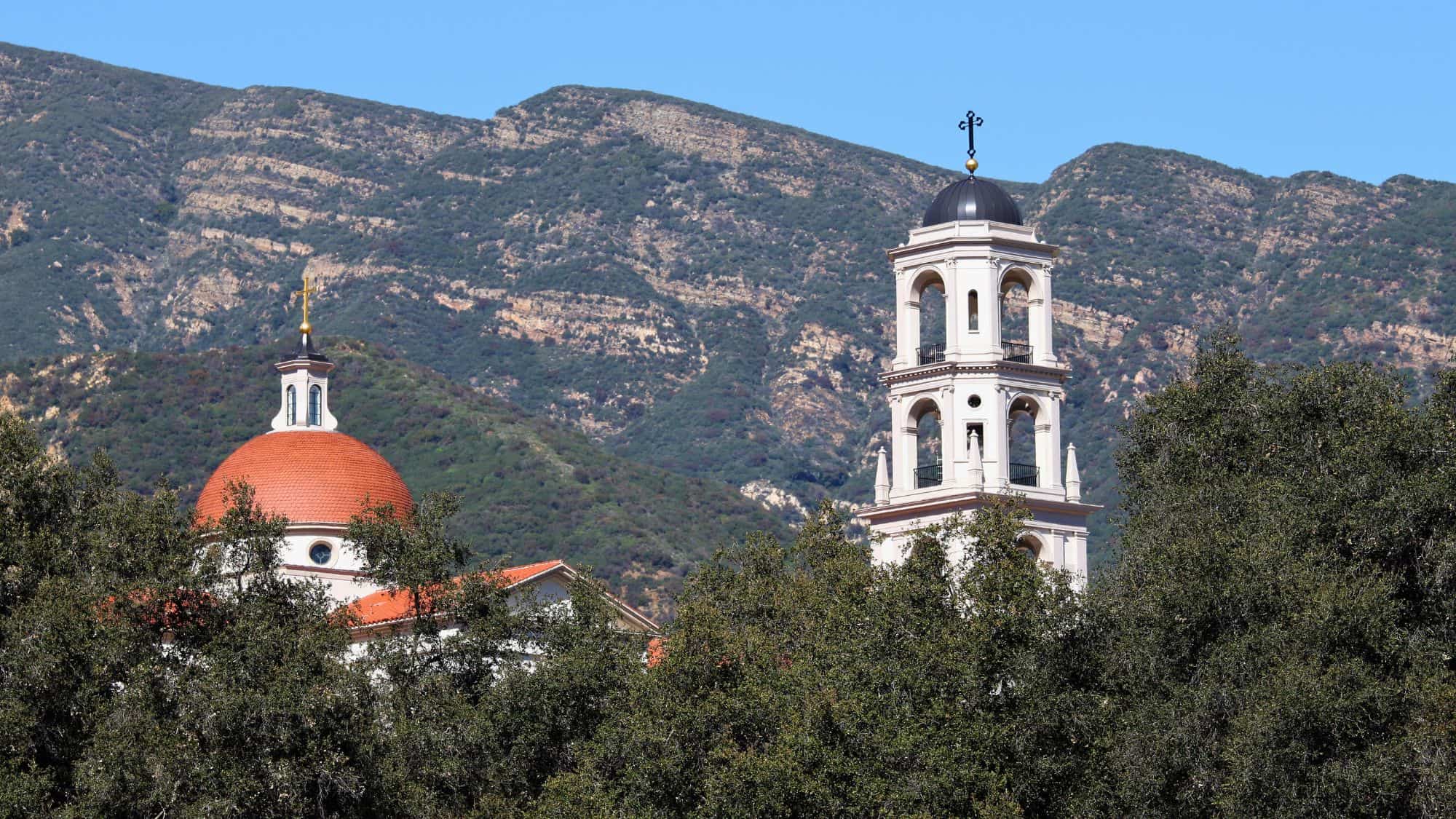 A red-tiled dome and a bell tower peek out from dense trees in front of California mountains, likely part of a mission-style building in the scenic town of Ojai.