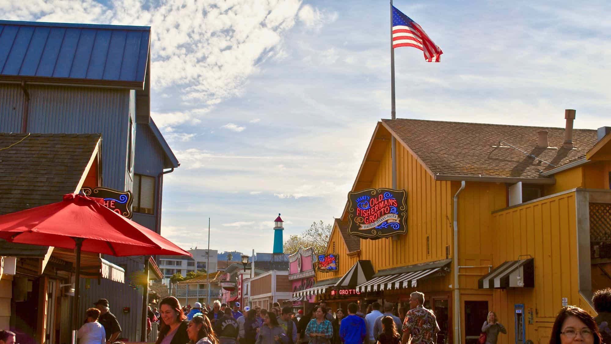 A bustling waterfront scene at Old Fisherman’s Grotto, a popular seafood restaurant in Monterey, California. Bright yellow buildings, a red umbrella, and a crowd of visitors create a lively atmosphere, with a lighthouse peeking out in the distance.