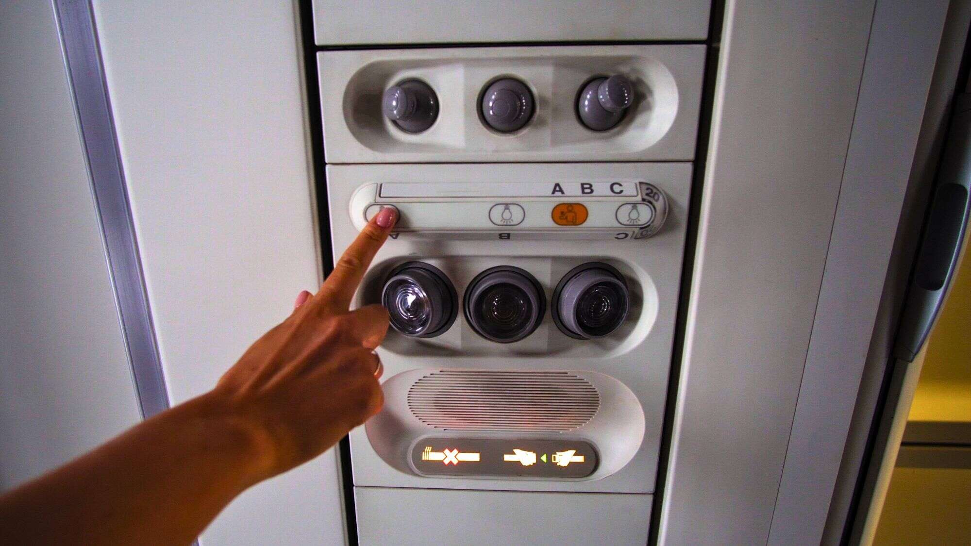 A hand reaches up to press a button above an airplane seat, surrounded by individual air vents, lights, and other control panel features.