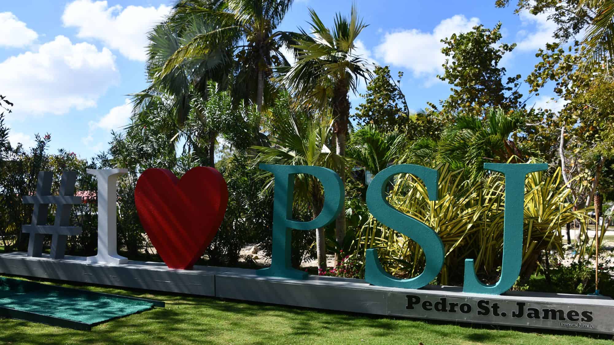 A lush garden surrounds a large outdoor sign that reads "#I ❤️ PSJ Pedro St. James," with tropical palm trees in the background. This historic site in the Cayman Islands offers a mix of heritage and scenic beauty.