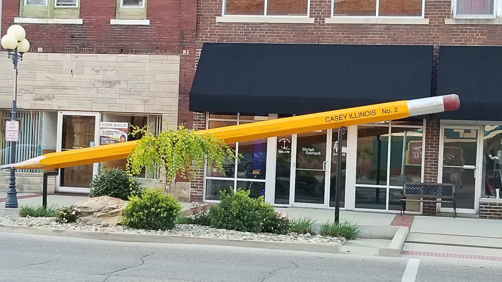 A massive yellow pencil sculpture, labeled “CASEY ILLINOIS No. 2,” is mounted diagonally in front of a brick building with black awnings.