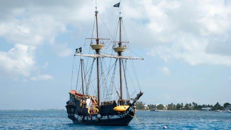 A classic tall ship with black pirate flags floats on the turquoise ocean, looking like a relic from the past. The ship is anchored near a tropical shoreline, giving off an adventurous, seafaring vibe.