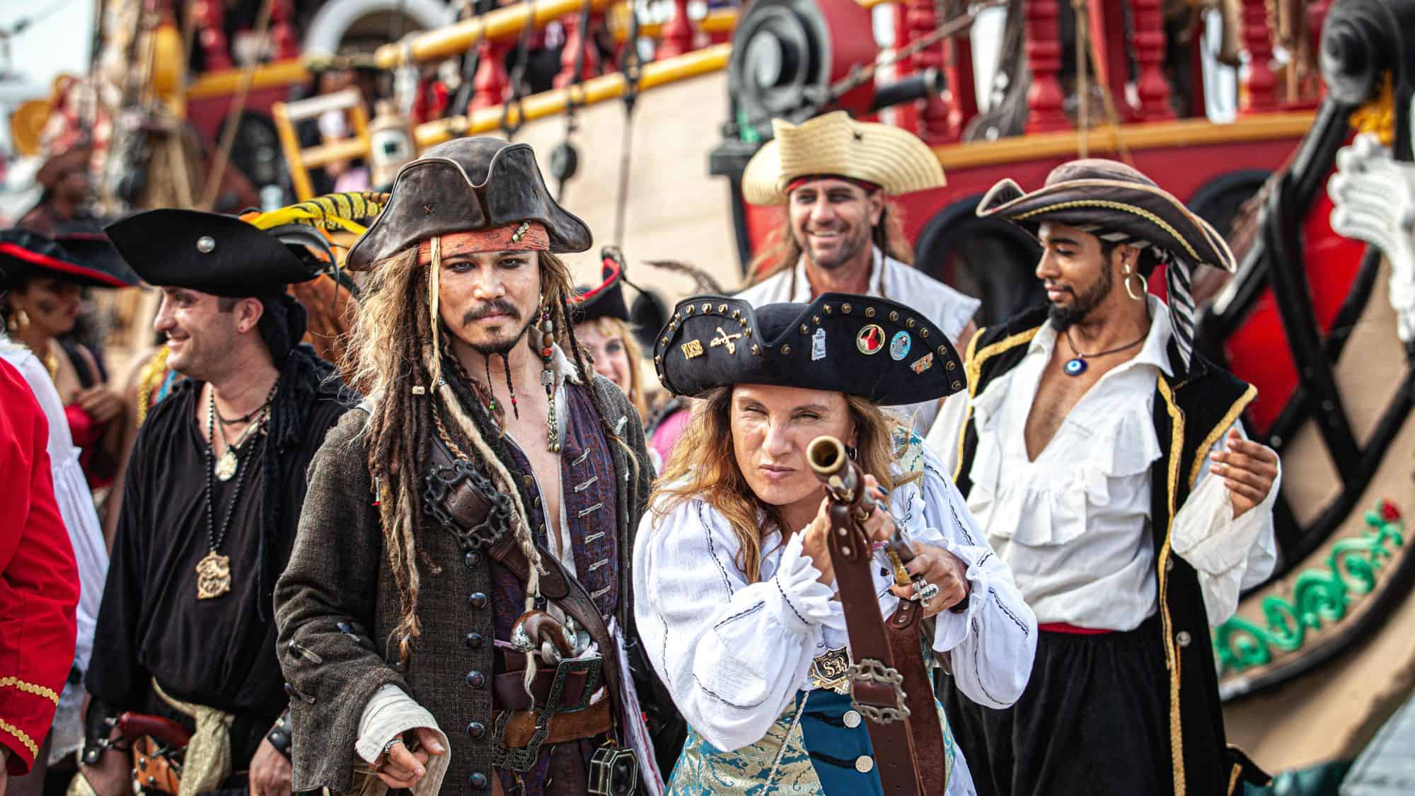 A lively group of people dressed as pirates parade through the streets, complete with tricorn hats, swords, and old-world attire. A woman in the foreground aims a small telescope, adding to the high-energy, swashbuckling scene.