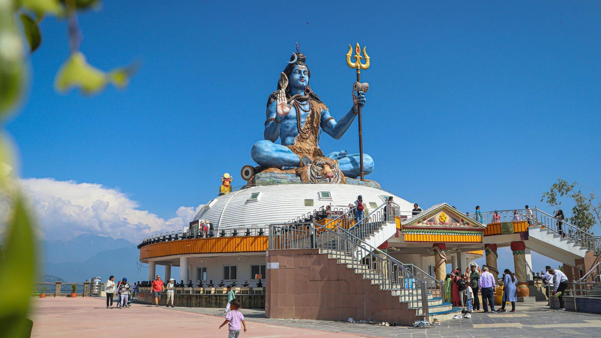 A massive blue statue of Lord Shiva sits atop a white temple dome in Pokhara, Nepal, with visitors walking around and a mountain range in the distance.
