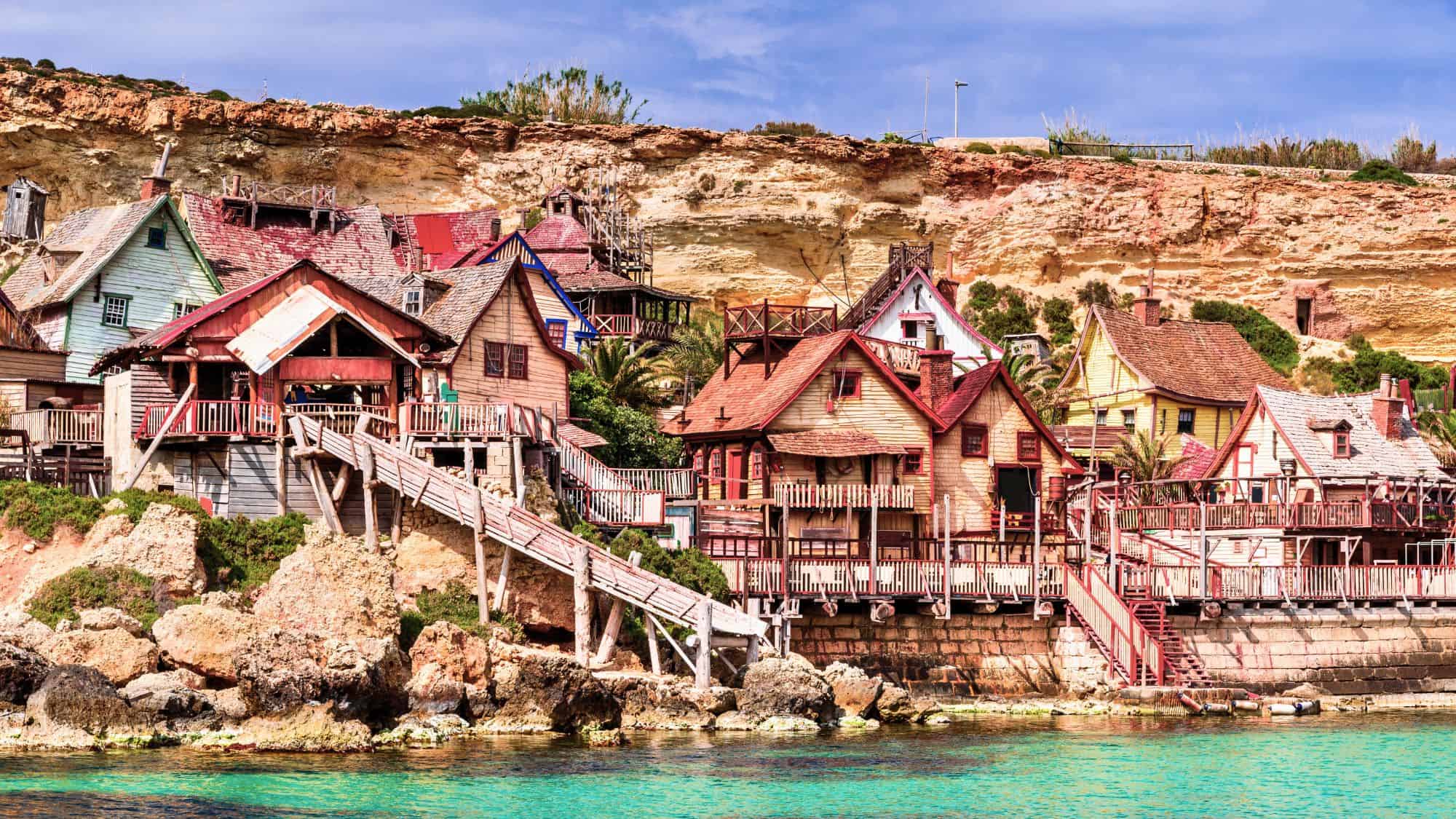 Colorful wooden buildings of Popeye Village clustered along the rocky Maltese coastline, with turquoise waters below and weathered cliffs behind, looking like a whimsical movie set.