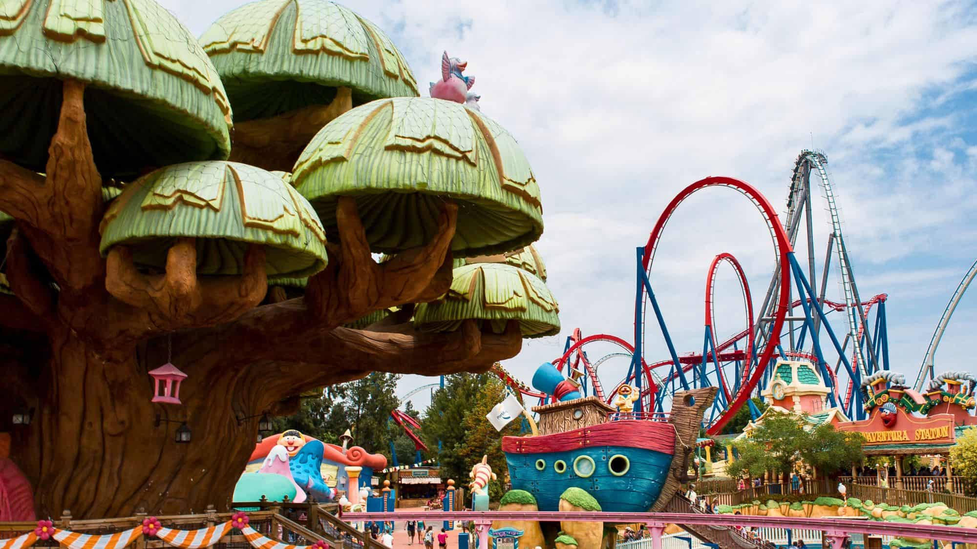 A whimsical fantasy-themed area at PortAventura Park in Spain, featuring oversized green mushroom structures, a colorful pirate ship, and towering red roller coasters.