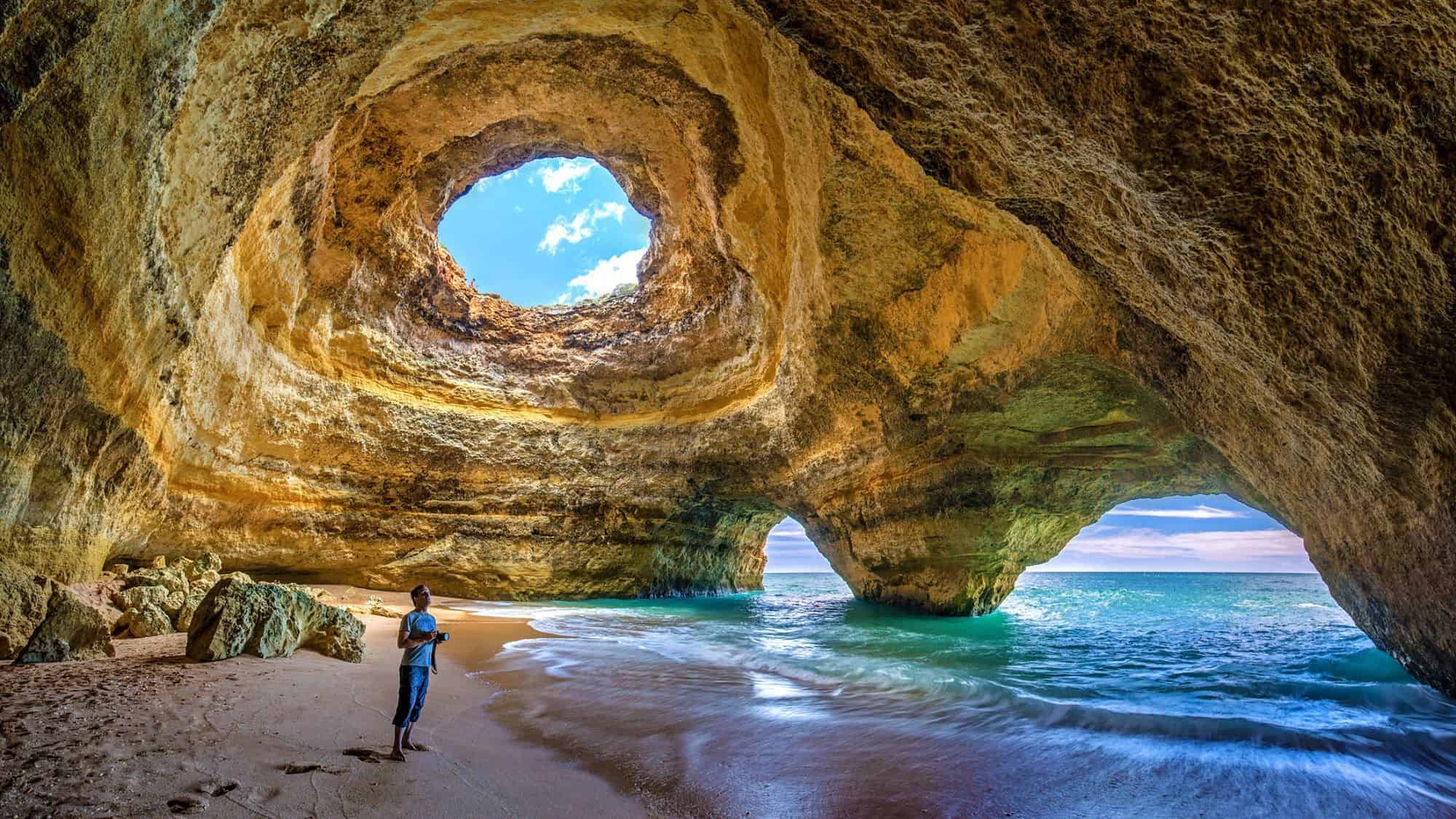 A picturesque shot of the Benagil Cave in Portugal, with sunlight streaming through the natural rock opening. The turquoise waves meet the sandy shore inside the cave, while a traveler admires the view.