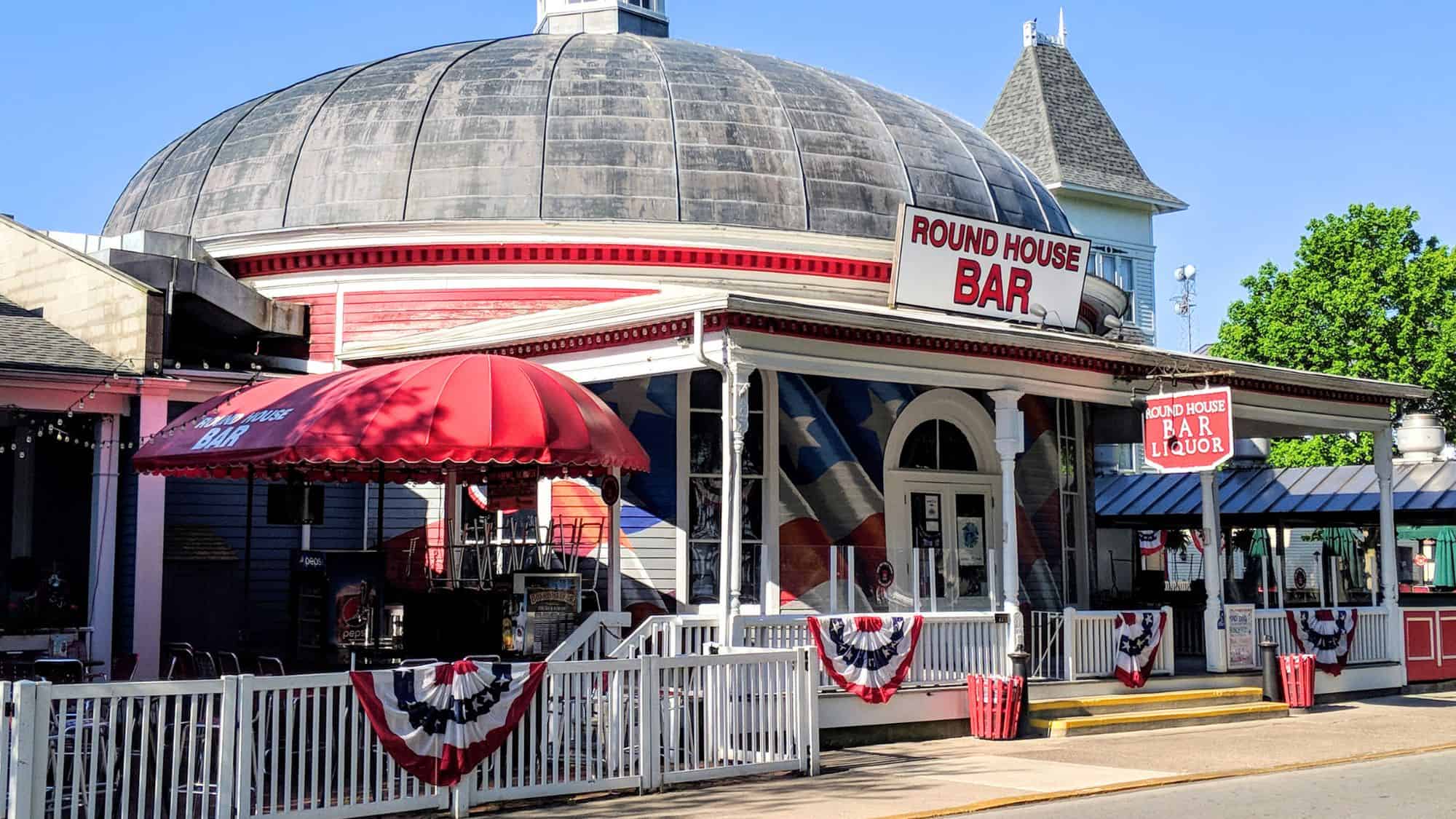 The historic Round House Bar with a red and white patriotic theme, featuring a domed roof and outdoor seating. American flags and bunting decorate the classic building, adding to its vintage charm.