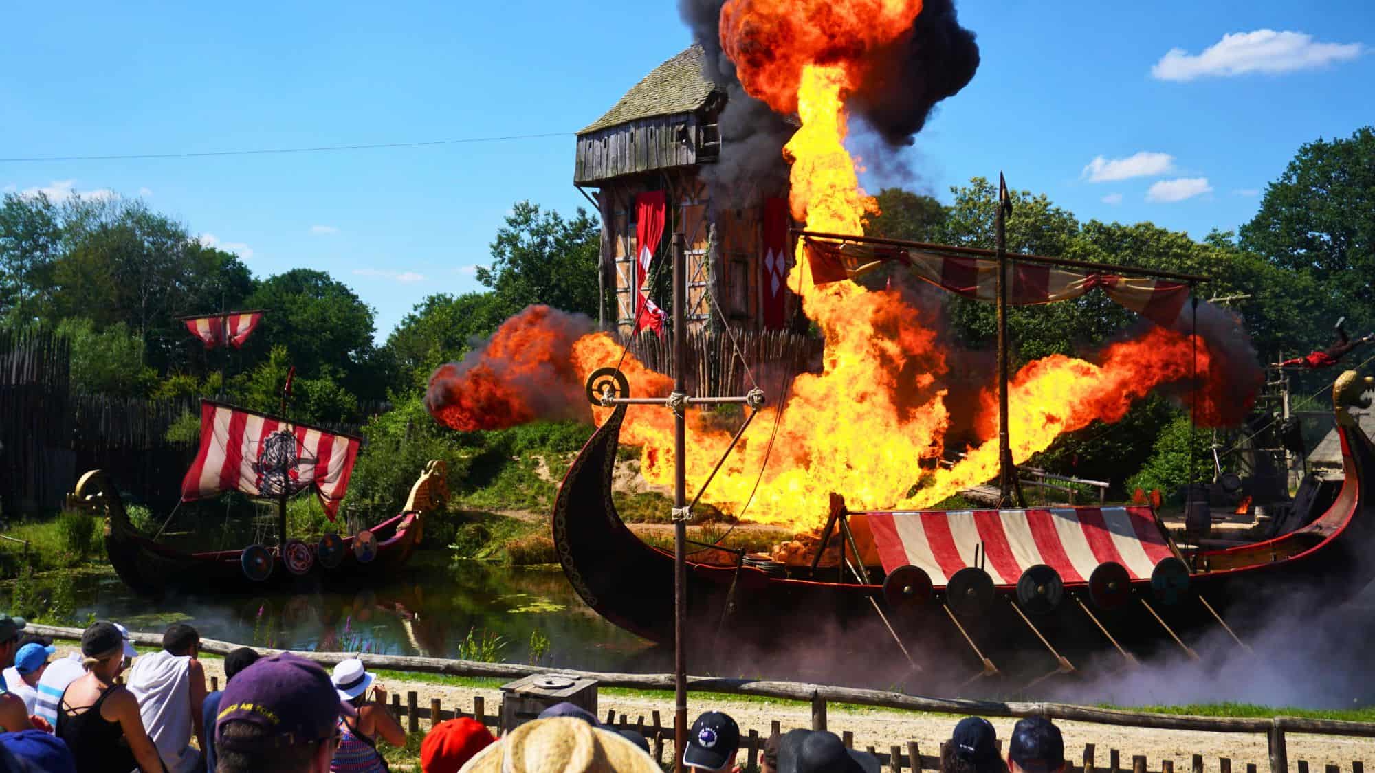 A dramatic stunt show at Puy du Fou in France, depicting a Viking ship under attack with flames and explosions, as an audience watches from the sidelines.