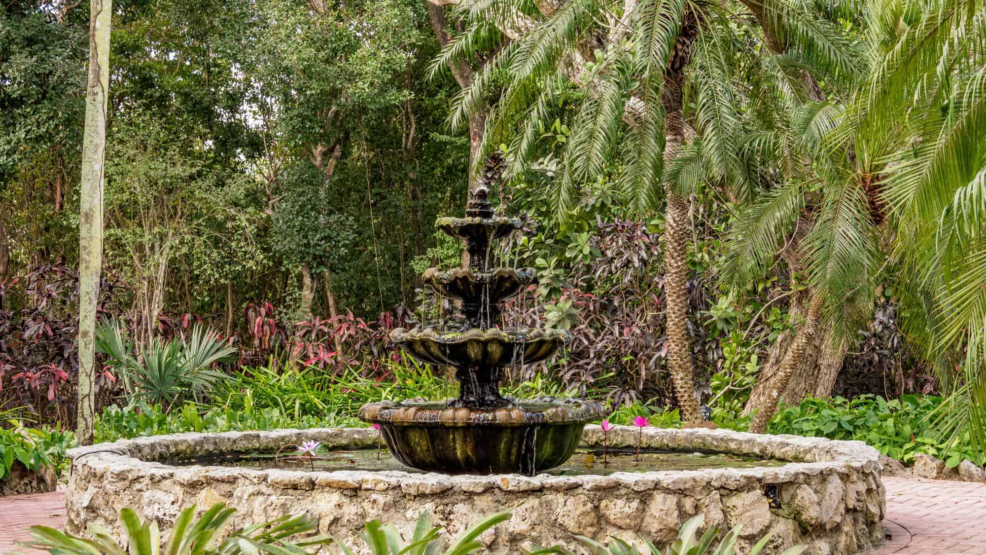 A tranquil stone fountain sits in the middle of a lush tropical garden, surrounded by palm trees and colorful plants. Water gently trickles from the fountain, enhancing the serene, natural setting.