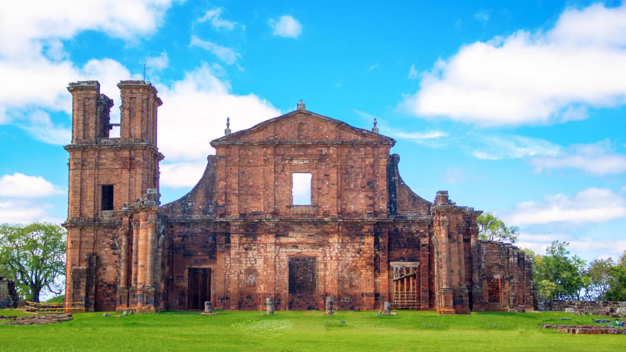 A historic Jesuit mission ruin in Paraguay, with a reddish-brick facade and two towering remnants of an old church. The lush green grass and bright blue sky contrast with the centuries-old structure.