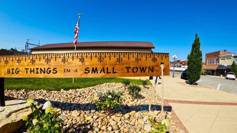 A wooden ruler sculpture with bold black measurement markings and the phrase “BIG THINGS IN A SMALL TOWN” stretches across a landscaped area in a small town.