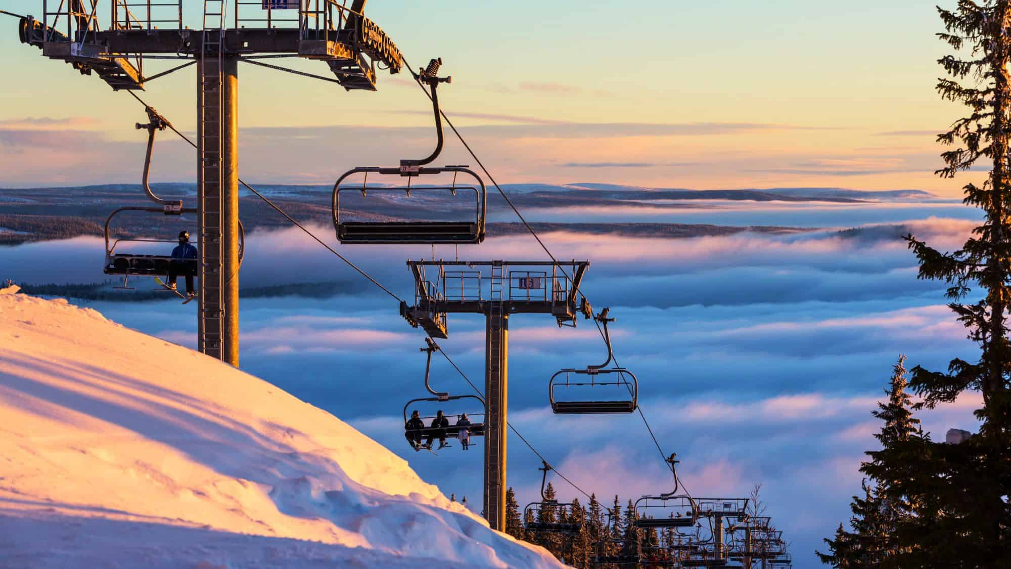 A ski lift ascending a snowy mountain at sunset, with layers of clouds below creating a dreamy, elevated feel. A lone skier is seen on the lift, enjoying the breathtaking view.
