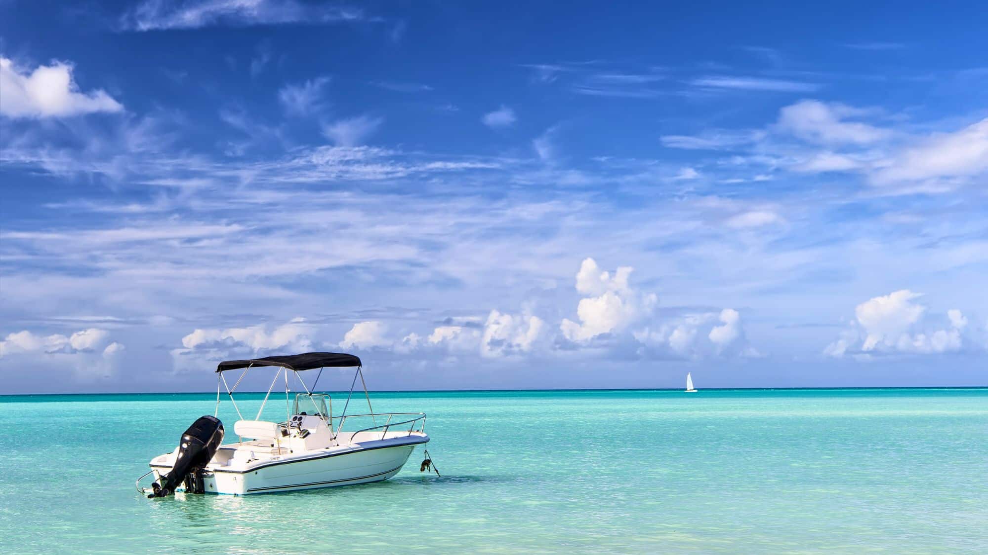 A small white motorboat floats on crystal-clear turquoise waters under a bright blue sky. A sailboat is visible in the distance, adding to the tropical paradise vibe.