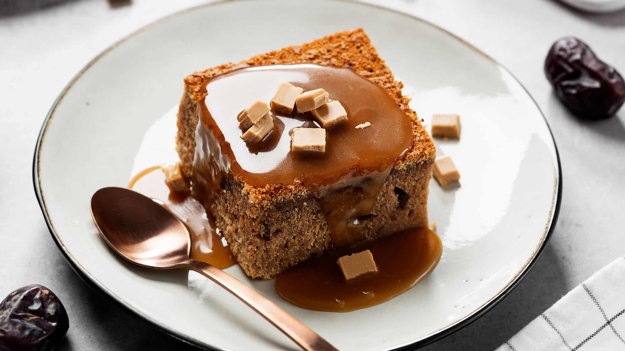 A moist square of sticky toffee pudding drizzled with warm caramel sauce and topped with small fudge pieces, served on a plate with a copper spoon and date fruit in the background.