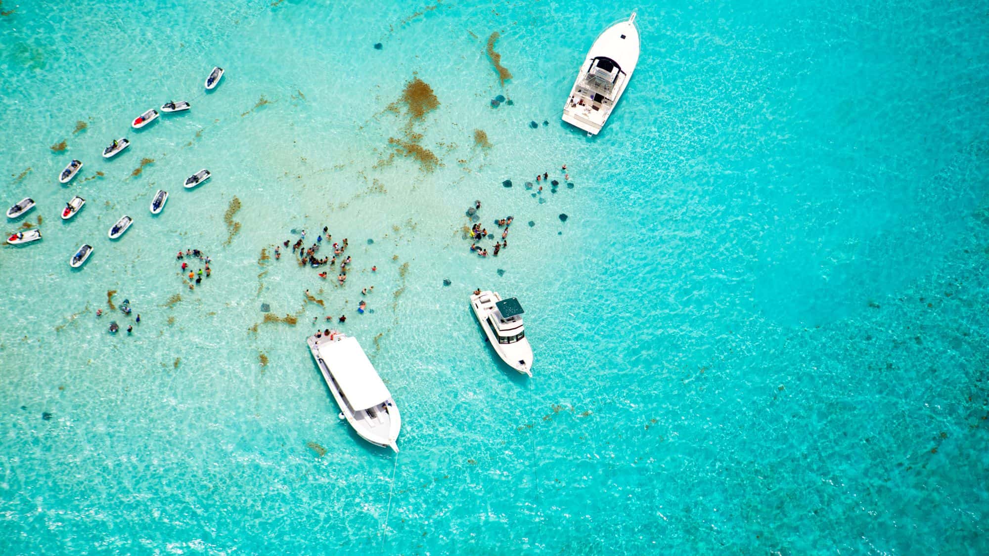 An aerial view of a shallow, crystal-clear turquoise sandbar where people are gathered, likely interacting with stingrays. Multiple boats and jet skis are anchored in the water, showcasing a popular Cayman Islands attraction.