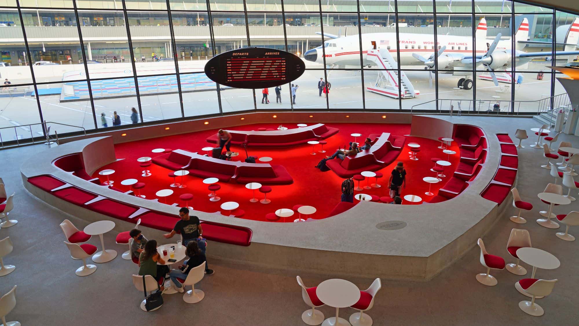 A retro-styled seating area with bold red carpet, curved benches, and white tulip tables, located in the TWA Hotel at JFK Airport, with large windows overlooking vintage planes outside.