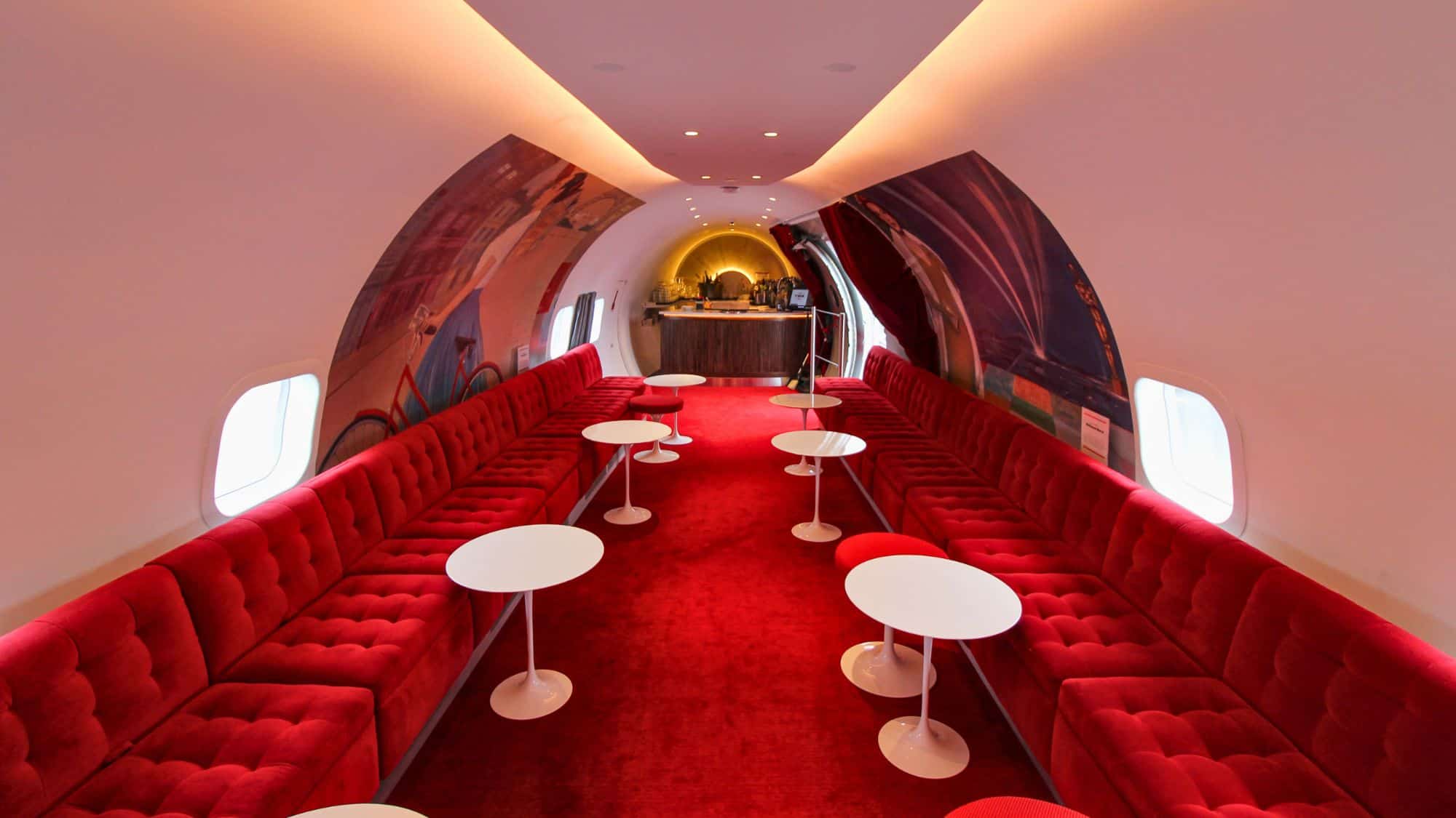 A stylish airplane lounge with red tufted seating, white tables, and a small bar at the far end, inside a repurposed aircraft, evoking a glamorous 1960s aesthetic.