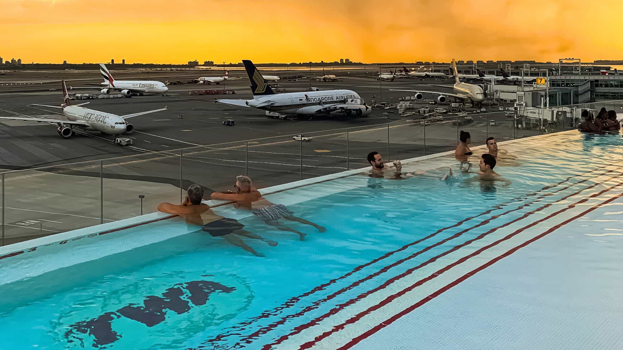 A rooftop infinity pool at the TWA Hotel, where guests relax while watching airplanes taxi and take off at JFK Airport, with a golden sunset in the background.